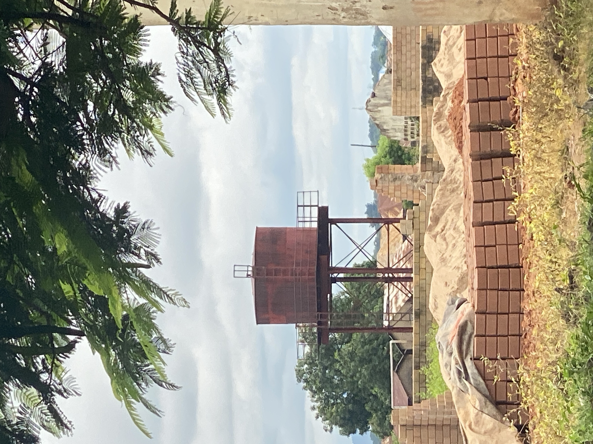 This is an outdoor, eye-level, medium shot of a water tower and construction materials in Bali, Nigeria. The sky is cloudy and overcast, with some trees visible in the foreground and background. In the foreground, there are stacks of bricks and piles of sand, covered in places with a tarp. Behind the construction materials, a large, rusty red water tower stands on a metal frame. Further in the background, several buildings, some incomplete, and lush green trees can be seen. The scene appears to depict a construction site in a semi-urban or rural area.