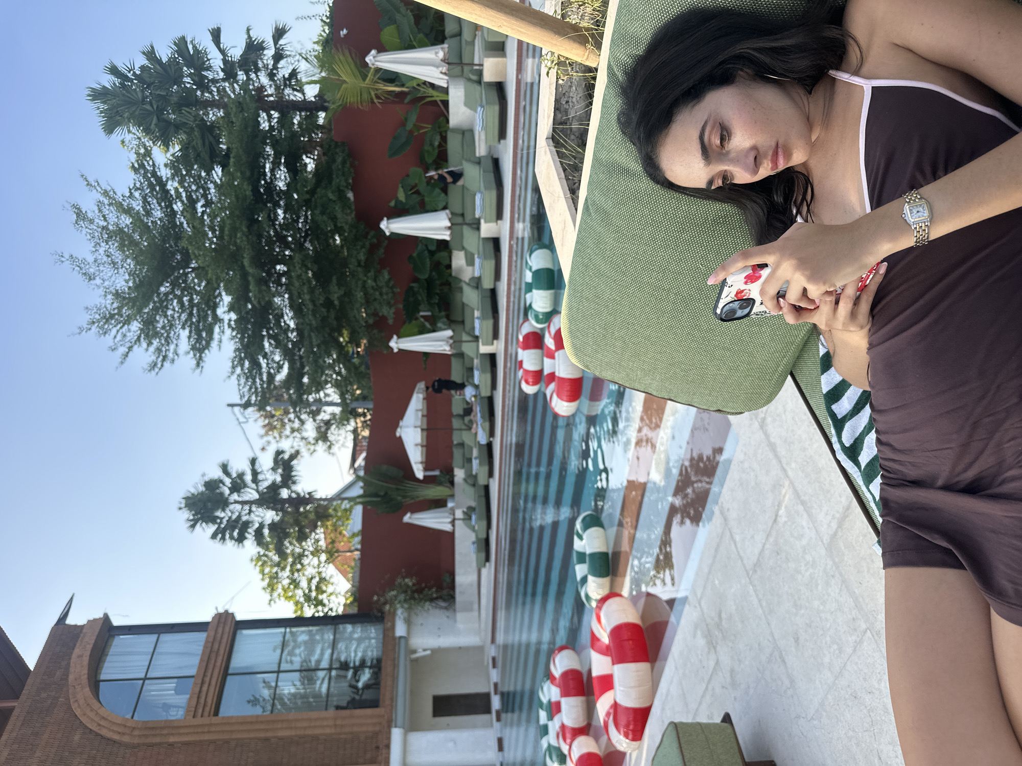 In an outdoor scene, a young woman with dark hair is reclining on a green lounge chair, looking at her phone. She is wearing a brown dress with a light pink trim. On her left wrist, she wears a silver watch with a white face. The lounge chair is positioned on a tiled patio next to a swimming pool filled with water and several red and green inflatable rings. In the background, there is a modern brick building with large windows, lush green trees, and palm trees. Several lounge chairs and white umbrellas are arranged around the pool. The sky is clear and blue.