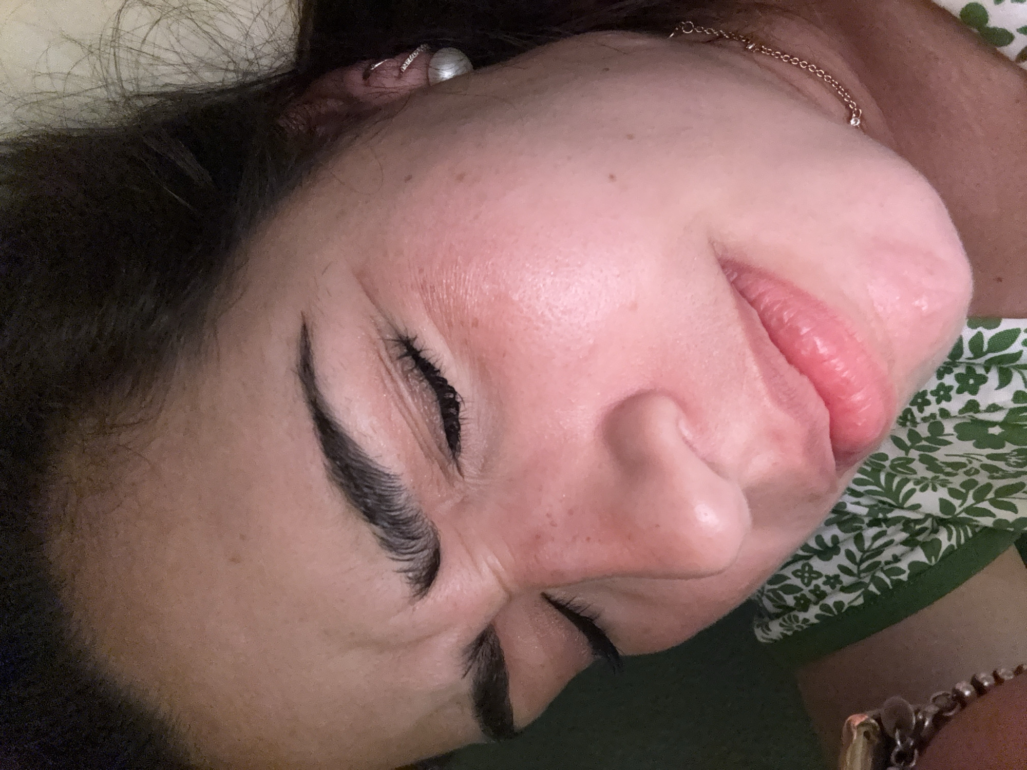 A close-up, slightly angled view shows a woman's face. She is lying down, with her head tilted slightly towards the right. Her eyes are closed, revealing long, dark eyelashes and defined eyebrows. The skin is fair with some freckles visible on her cheek. A pearl earring is seen in her left ear. A gold necklace sits on her neck. A green and white floral pattern can be seen near her collar. The background appears to be dark green. The location is most likely indoors, in the United States of America. The city is not discernible from the image.