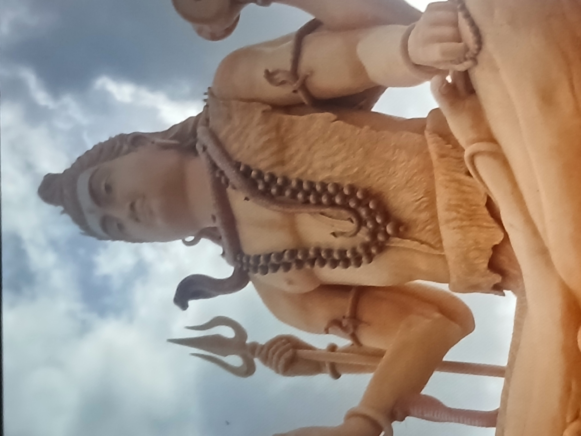 The image shows a statue of the Hindu god Shiva. The statue is light tan in color and is holding a trident. The statue is wearing a rudraksha mala necklace. In the background is a cloudy sky. The statue is located in Bengaluru, India.