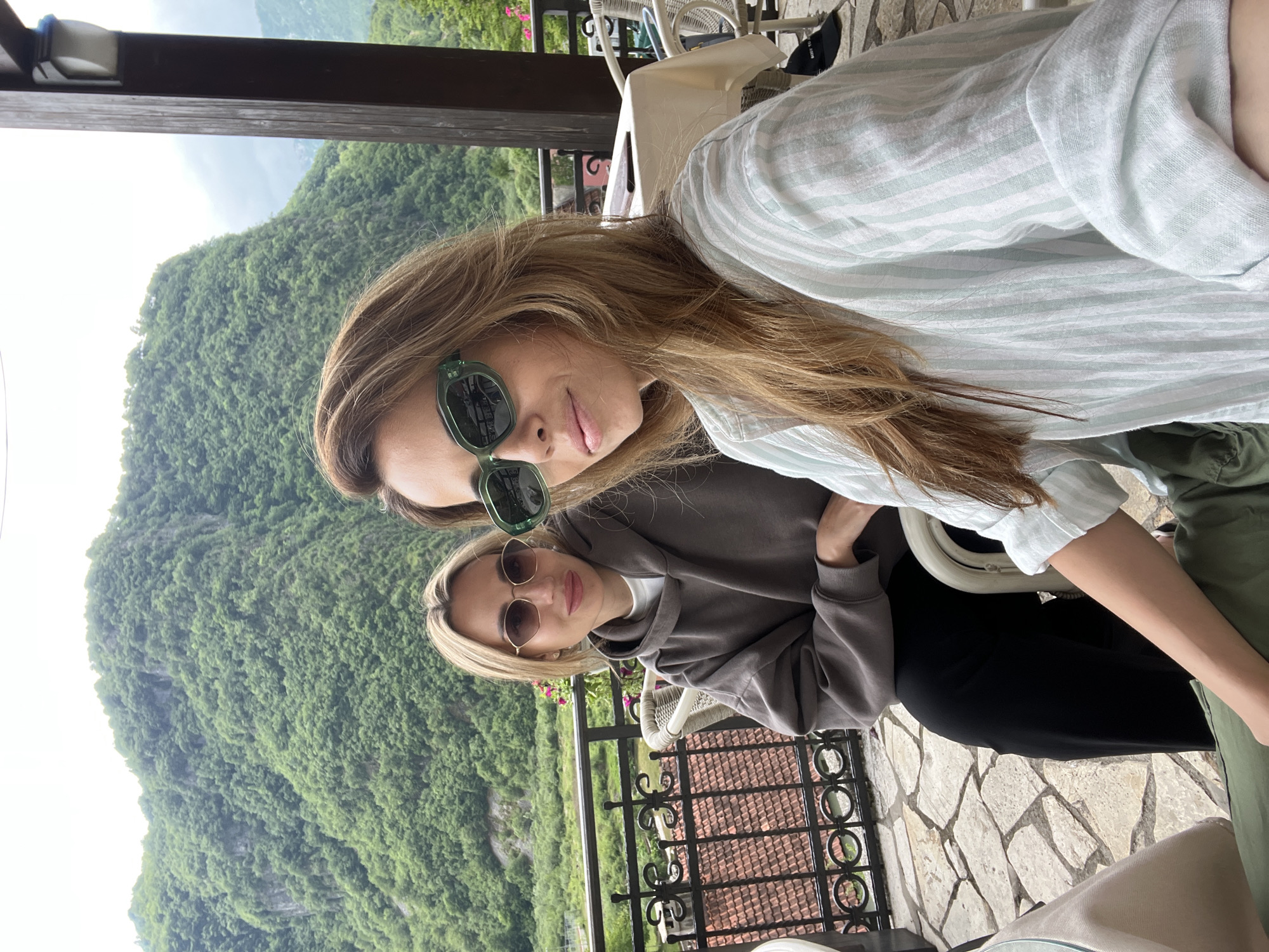This outdoor portrait features two women on a balcony or terrace, overlooking a picturesque mountainous landscape, likely in Šavnik, Montenegro. The scene is set during the daytime under an overcast or hazy sky.
The woman in the foreground, with long, light brown hair, wears dark green sunglasses and a light blue and white striped long-sleeved shirt, displaying a relaxed, slight smile. Behind her and slightly to her right, another woman with blonde hair is seated, wearing light-colored, round sunglasses, a dark grey hooded sweatshirt over a white shirt, and black pants. Both women appear calm and are posing for the camera.
The background showcases a magnificent, large mountain covered in dense green foliage, emphasizing a lush, natural environment. Below the ornate black metal railing of the balcony, glimpses of red-tiled roofs are visible, suggesting a settlement or village nestled in the valley. The terrace floor is paved with irregular, light-colored stone tiles, and white wicker-style chairs and tables are present, indicating a cafe or restaurant setting. No discernible text is visible in the image. The overall atmosphere conveys tranquility and appreciation for the scenic surroundings.