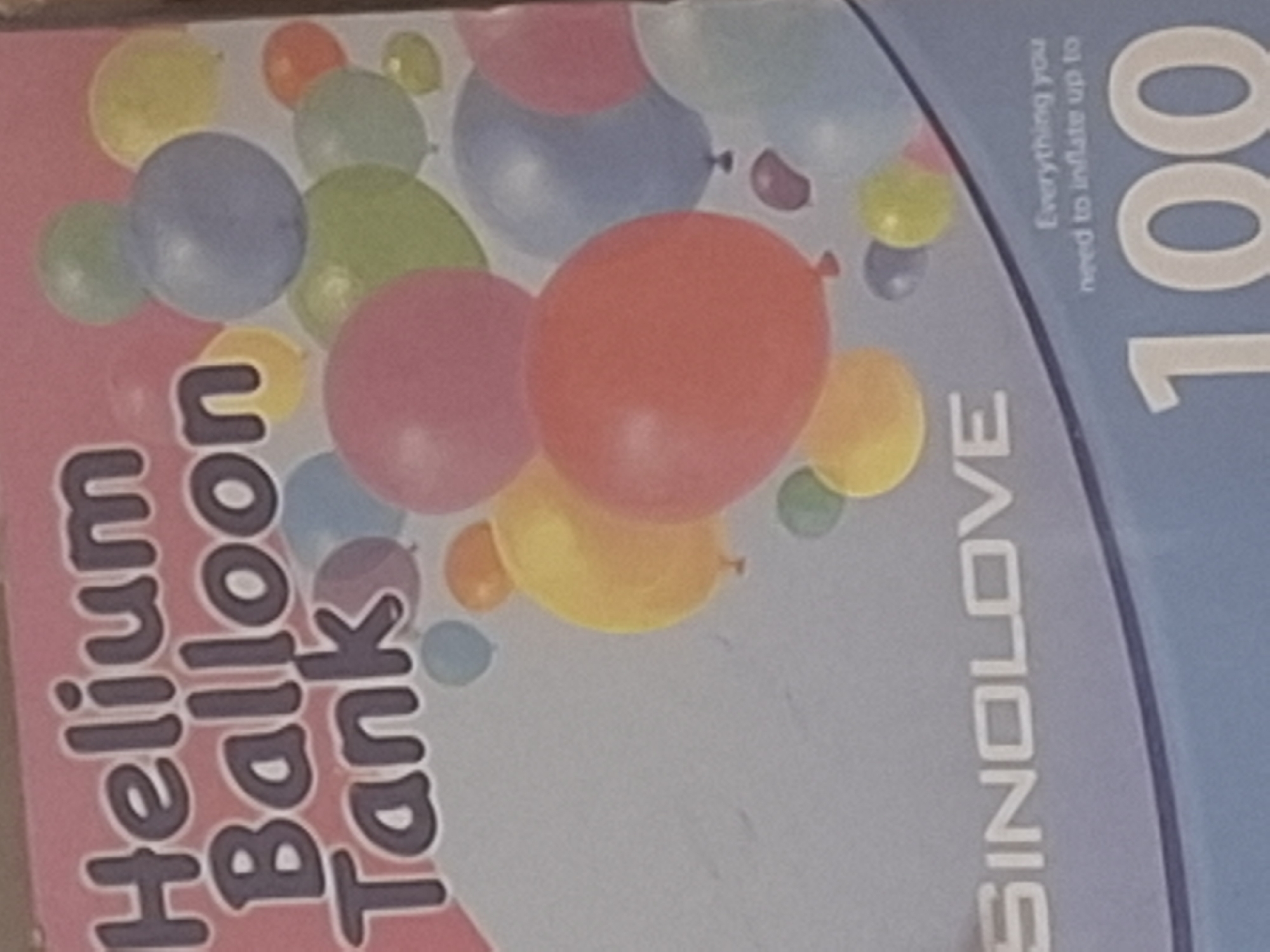 The image is a close-up, slightly blurry photograph of a pink and blue box that appears to be packaging for a helium balloon tank. The box has text that reads "Helium Balloon Tank" in large, dark purple, stylized letters. Below this, in white, is the brand name "SINOLOVE". Scattered across the box are various colorful balloons in shades of pink, blue, green, yellow, and orange. Towards the bottom right, there is smaller text that says "Everything you need to inflate up to" followed by a large "100". The background of the box is a gradient of light pink at the top to a light blue at the bottom. There is no visible indication of the city of Mıñbaev, Kazakhstan, in the image. The photograph is taken at a slight angle, suggesting it might be a product photo or a shelf display.