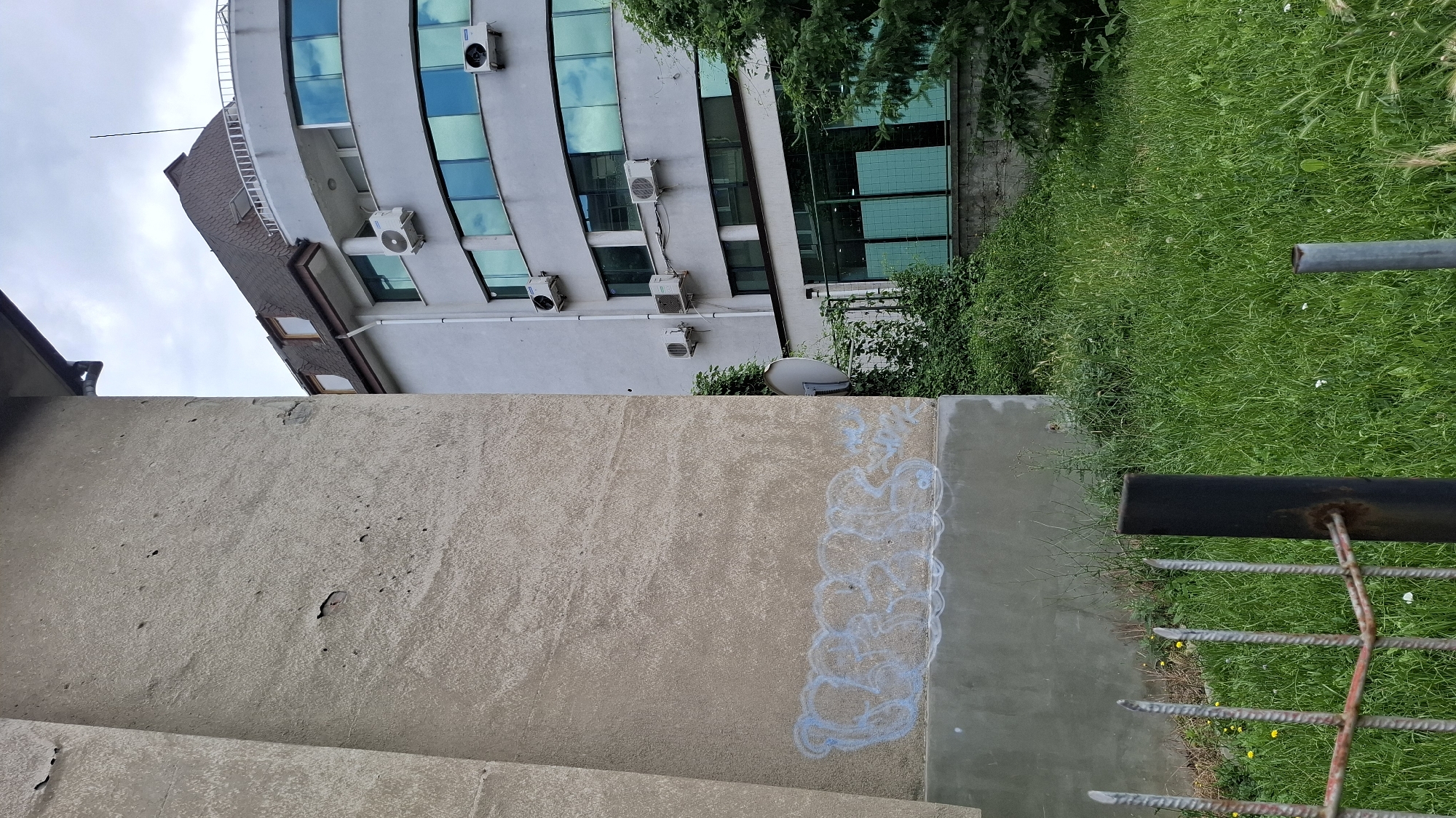 A low-angle, close-up, outdoor shot shows a weathered, beige concrete wall on the left, partially covered with light blue graffiti that is faded and illegible. To the right of the wall, a modern, multi-story building with a curved facade and large, reflective blue-tinted windows is visible. The building has several outdoor air conditioning units mounted on its exterior. A dark, sloped roof with dormer windows caps the building. Lush green grass and dense vegetation grow in the foreground and around the base of the building, obscuring a fence made of rusty metal bars and dark metal posts. The sky is overcast with gray clouds. The scene appears to be located in an urban environment, possibly Sofia, Bulgaria, given the architectural style and urban decay elements.
