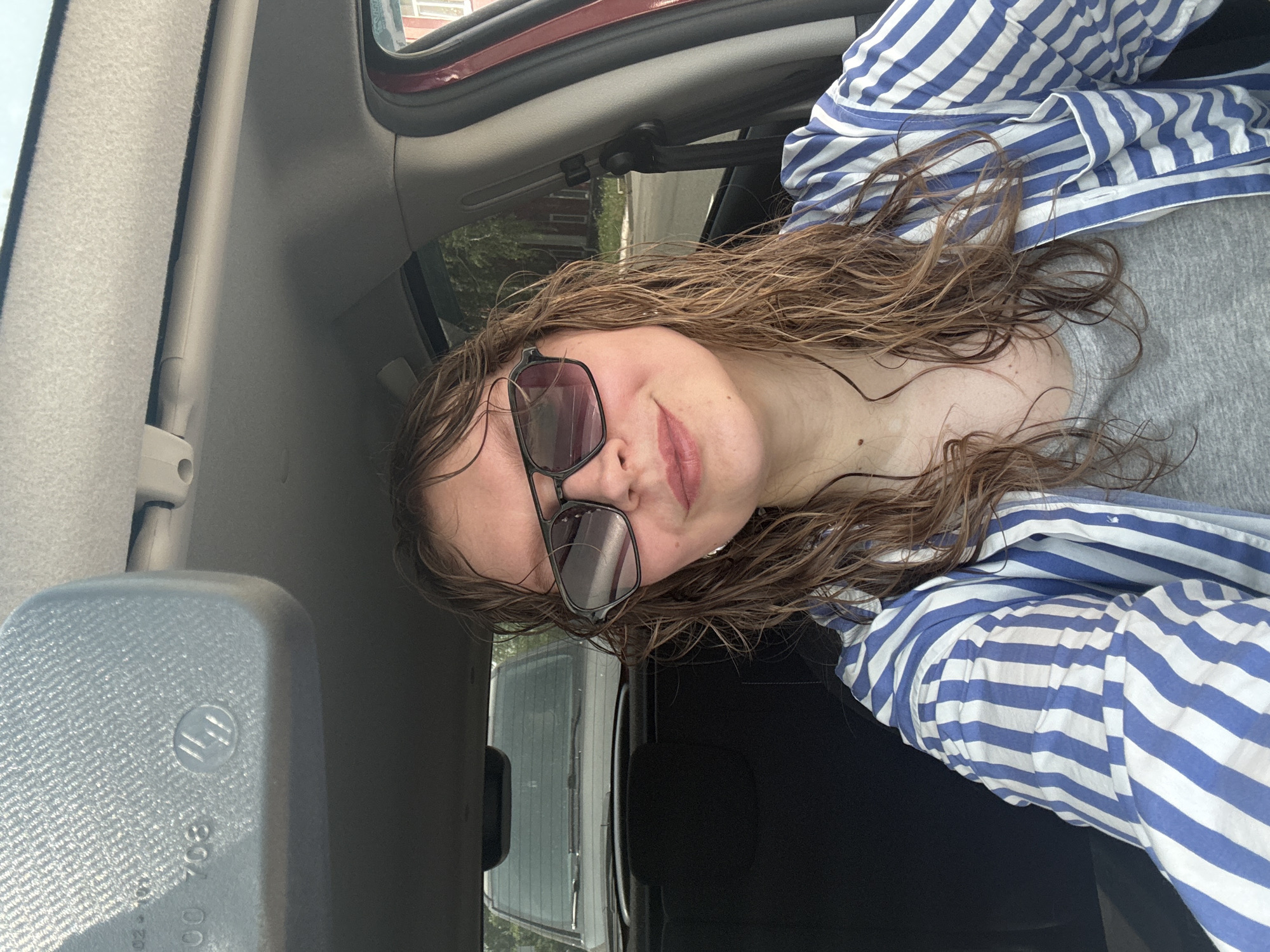 A woman with wet, brown curly hair and sunglasses sits in the passenger seat of a vehicle. She wears a grey shirt and a blue and white striped button-down shirt. The vehicle's interior is visible, including the headliner, door frame, and rearview mirror. Outside the vehicle, trees and buildings are visible, suggesting an urban environment. There is no specific information present to identify the location as Izhevsk, Russia.