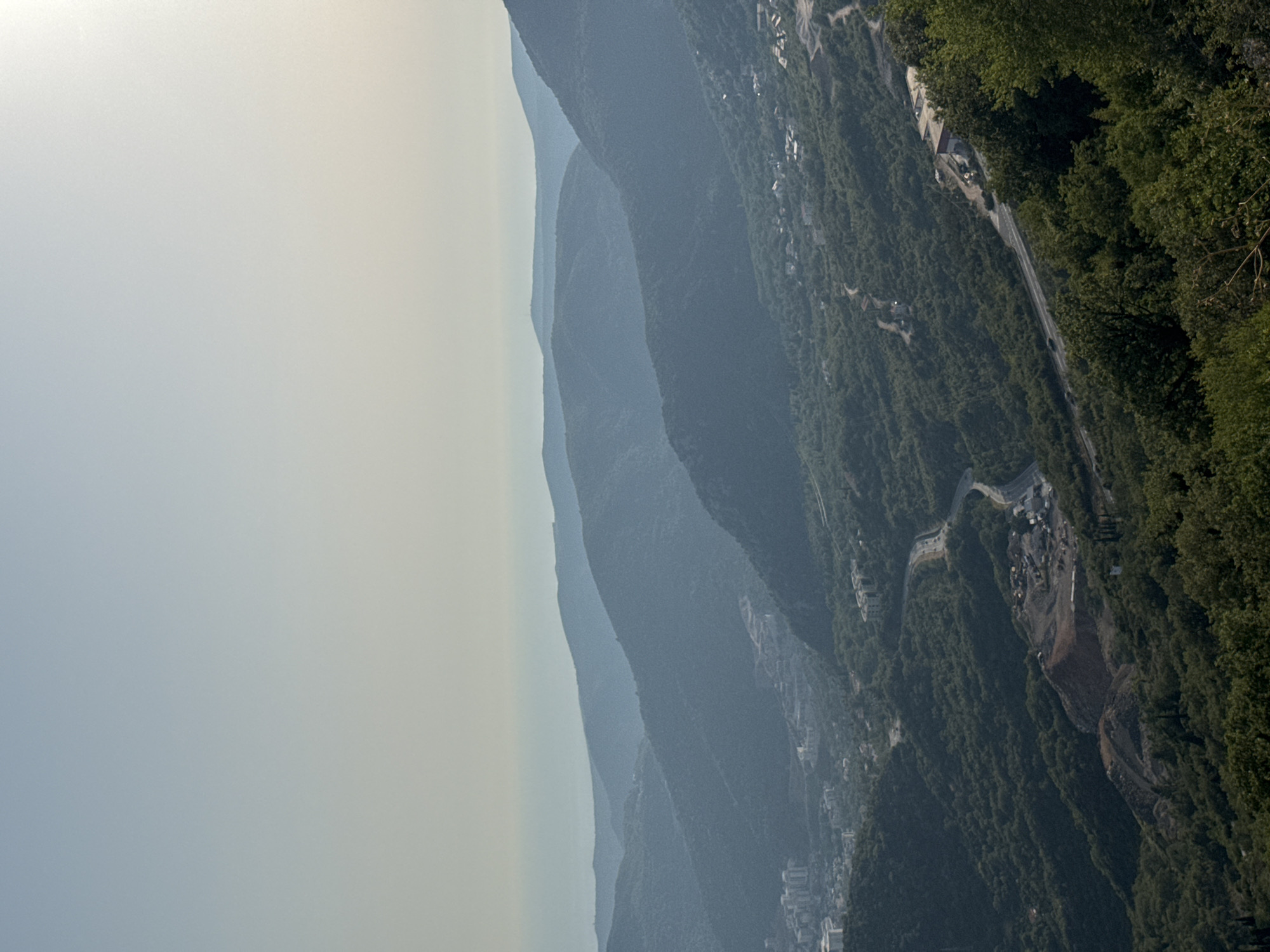 This is an aerial, wide-angle, daylight photograph of a mountainous landscape in Brajici, Montenegro. The scene is dominated by layers of blue-hued mountains receding into the distance, with a hazy horizon where the sea meets the sky. The foreground is a dense, green forest that covers the slopes of the mountains. A winding road is visible snaking its way through the trees, with a few buildings and a construction site appearing on the hillsides. The sky is pale blue and appears to be overcast or hazy. There are no people or animals visible in the image. The overall impression is one of a vast, undeveloped natural landscape, with human presence indicated by the road and construction.