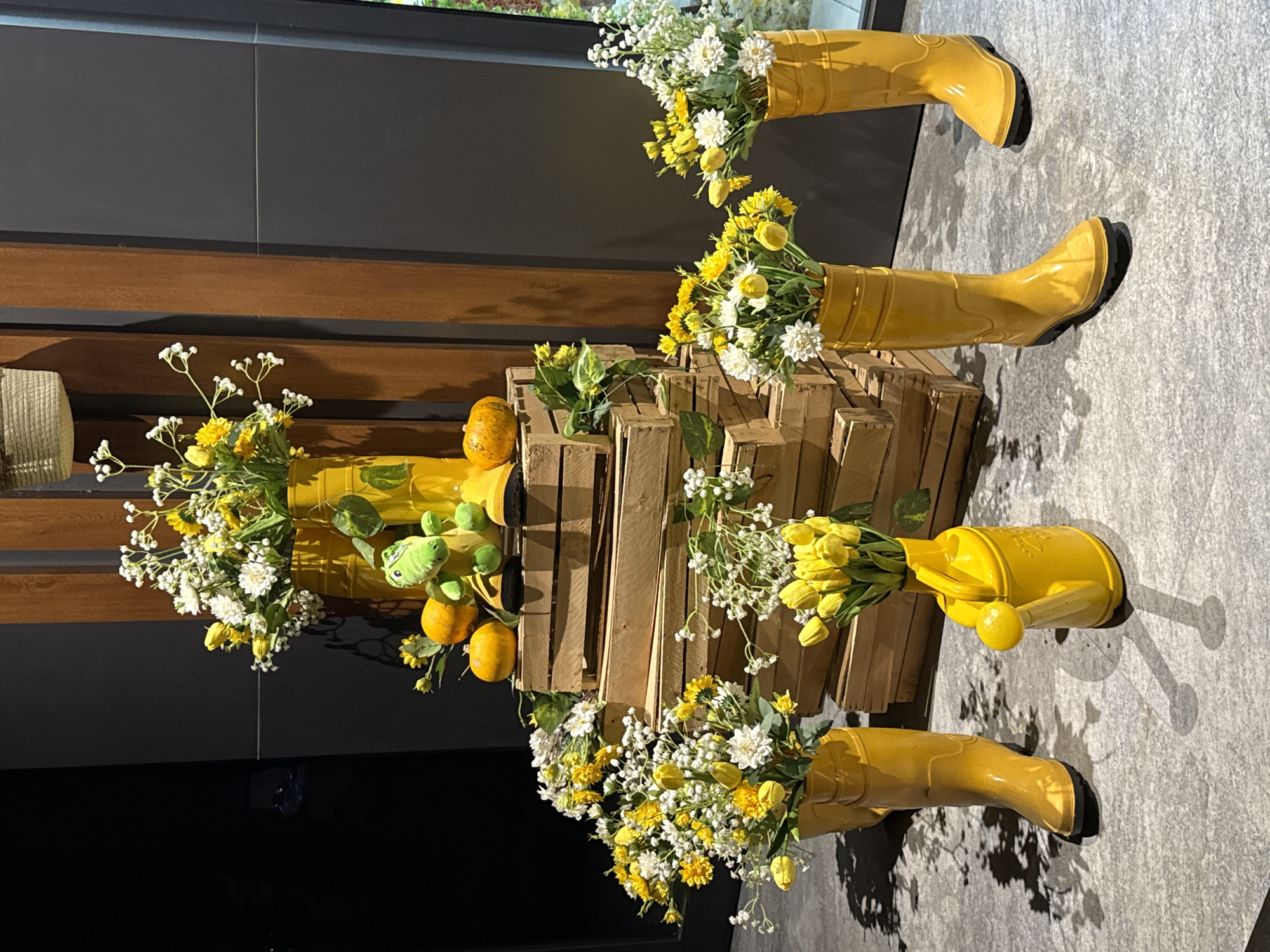 This is a well-lit, eye-level, medium shot of a decorative display. The scene is likely in Tashkent, Uzbekistan, given the implied context from the prompt. The display features a rustic theme with yellow accents.
In the background, a dark wall is visible to the left, and to the right, a tall, vertical wooden element with a textured surface, possibly a decorative panel or structural beam. A woven basket is suspended from above and slightly to the left of the wooden element.
The central focus of the image is a tiered arrangement of stacked wooden crates, creating a stepped platform. On top of these crates, a green plush toy dinosaur is seated between two small, orange pumpkins. The dinosaur is wearing a pair of yellow boots.
Several arrangements of yellow and white flowers are displayed prominently throughout the scene. To the left, a bouquet of yellow and white flowers is in a yellow Wellington boot, and next to it, another yellow Wellington boot is filled with yellow tulips and white filler flowers. In the foreground, to the right of the boots, a yellow watering can is filled with yellow tulips. Further to the right, another yellow Wellington boot is filled with yellow and white flowers.
The floor appears to be a textured gray surface, and there are dappled shadows cast by the objects, suggesting overhead lighting. The overall impression is a cheerful and whimsical display, possibly for a seasonal decoration. There are no people visible in the image.