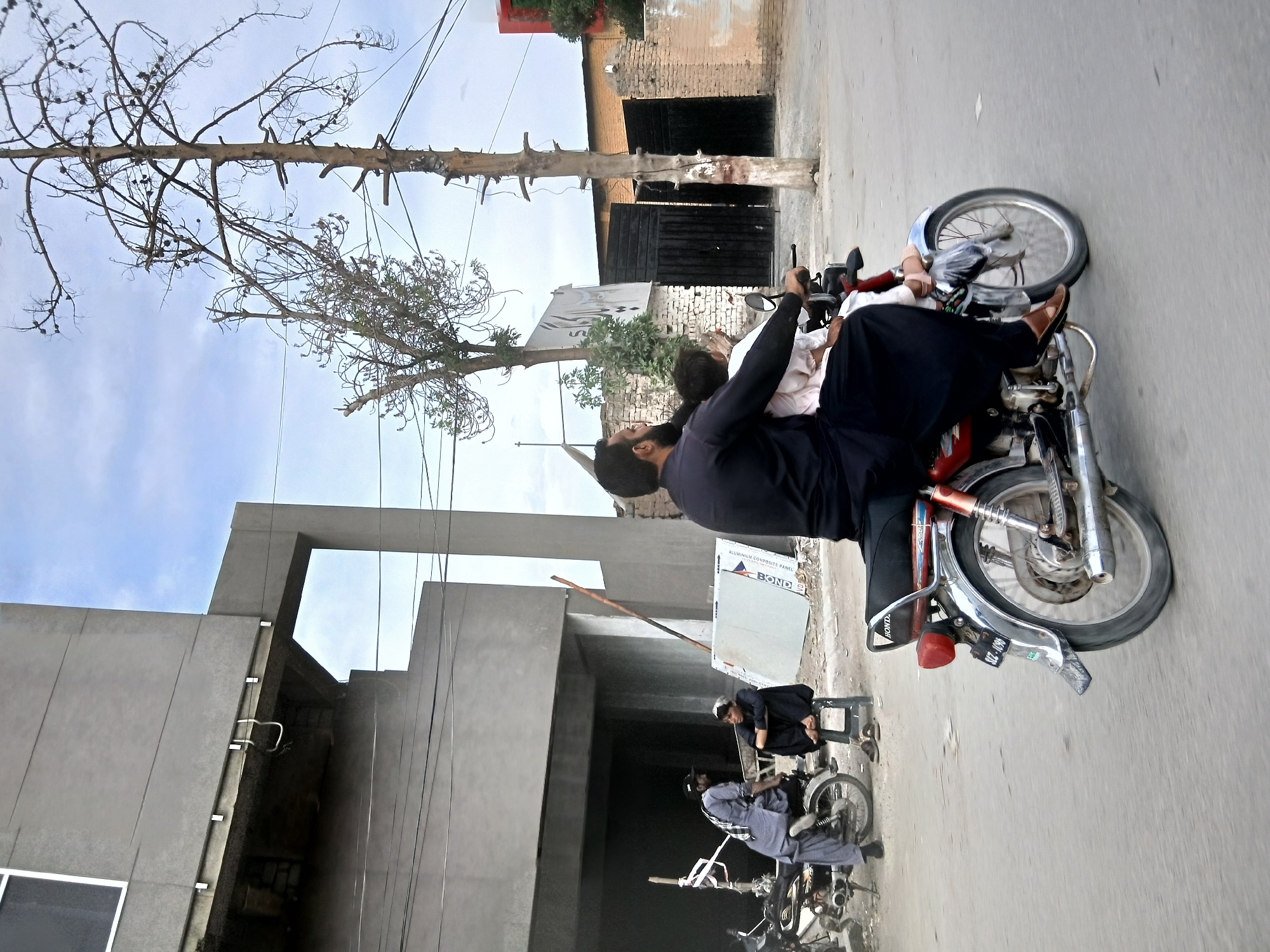 A street scene in Quetta City Tehsil, Pakistan shows a road with at least three motorcycles. One motorcycle has two men in dark clothing; the driver wears brown shoes, and the passenger is wearing what looks like white or off-white shoes. In the background there is a building, and a tree. In the bottom right there is another motorcycle with a man who is looking down at his phone. Further back, a third motorcycle with a man in grey clothes can be seen.