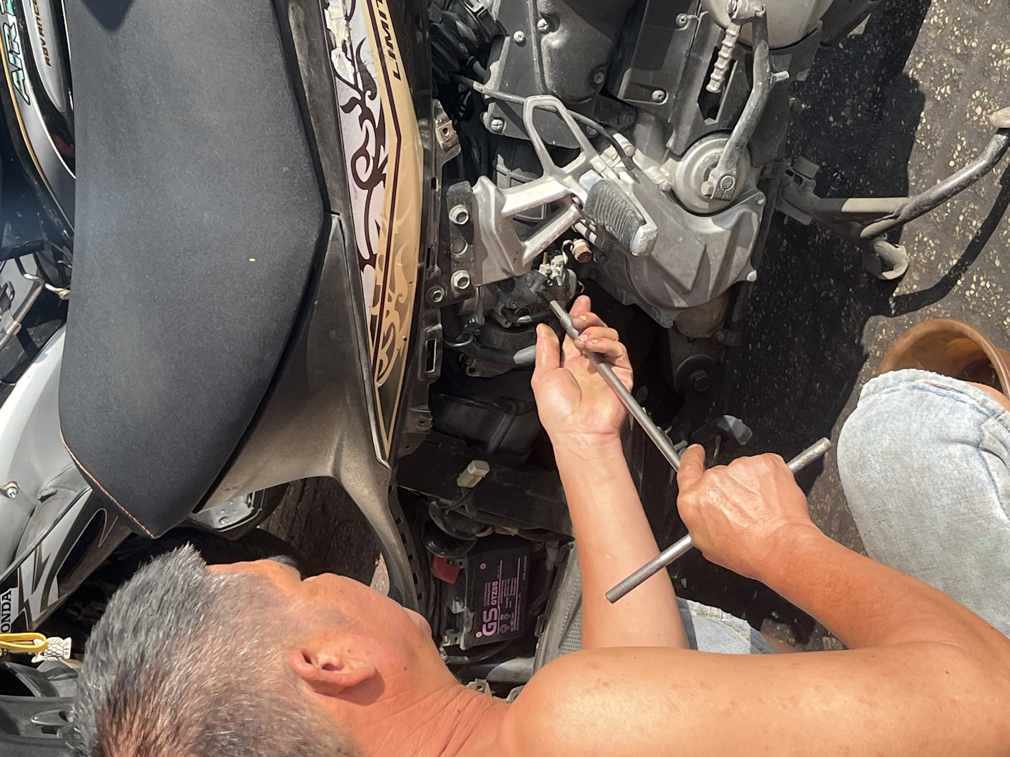 A man is working on a motorcycle in Bac Nha Trang Ward, Vietnam. The man is positioned close to the motorcycle, using a pair of wrenches to work on the engine area. The motorcycle is a light-colored model with a black seat. The setting is outdoors on a paved surface. The man appears to be shirtless. The scene is lit by natural daylight.