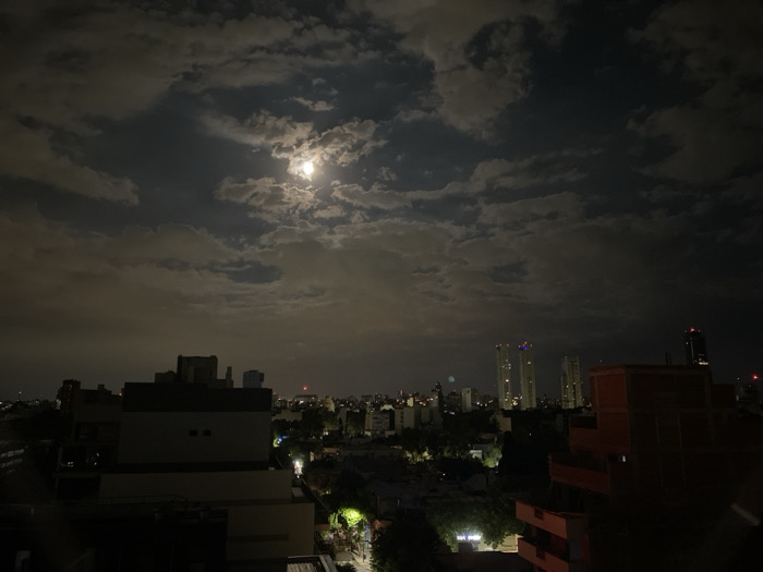 A night cityscape dominated by a cloudy sky with a bright moon peeking through. City buildings and streetlights are visible in the lower portion of the image. Some taller structures can be discerned in the distance, and the closest buildings appear silhouetted. The overall atmosphere is dark with patches of diffused light.
