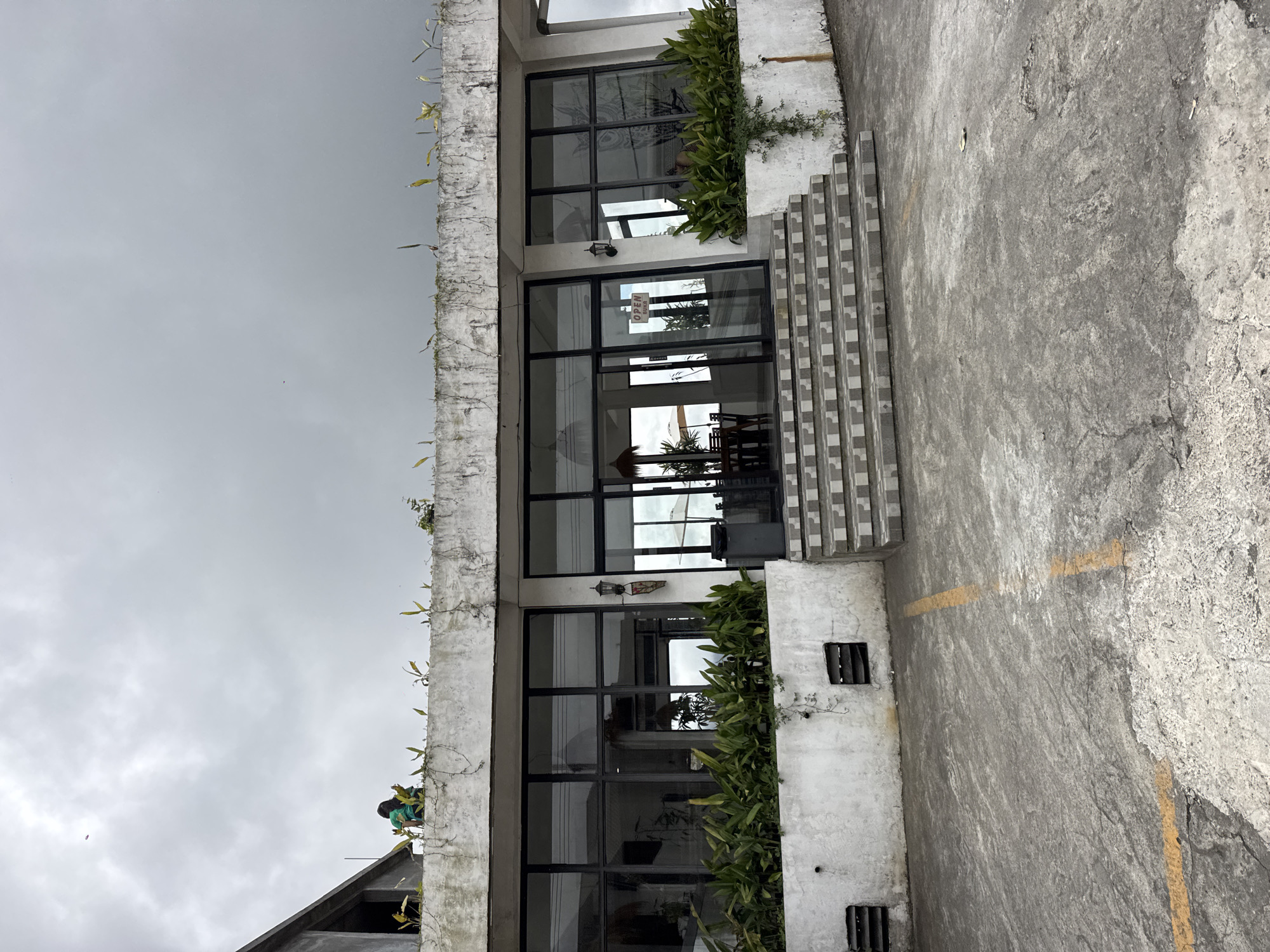 In Kintamani, Indonesia, a two-story building with large glass windows and a white concrete facade stands under a cloudy sky. The building's upper level features a terrace with sparse vegetation and two individuals, one wearing a green shirt, visible near the edge. The lower level has a prominent set of tiled stairs leading to glass doors. Lush green plants line the base of the building. A gray trash can is positioned to the right of the stairs, and ventilation grates are set into the lower wall. The foreground consists of a cracked concrete surface with yellow painted lines, suggesting a parking area or pathway.