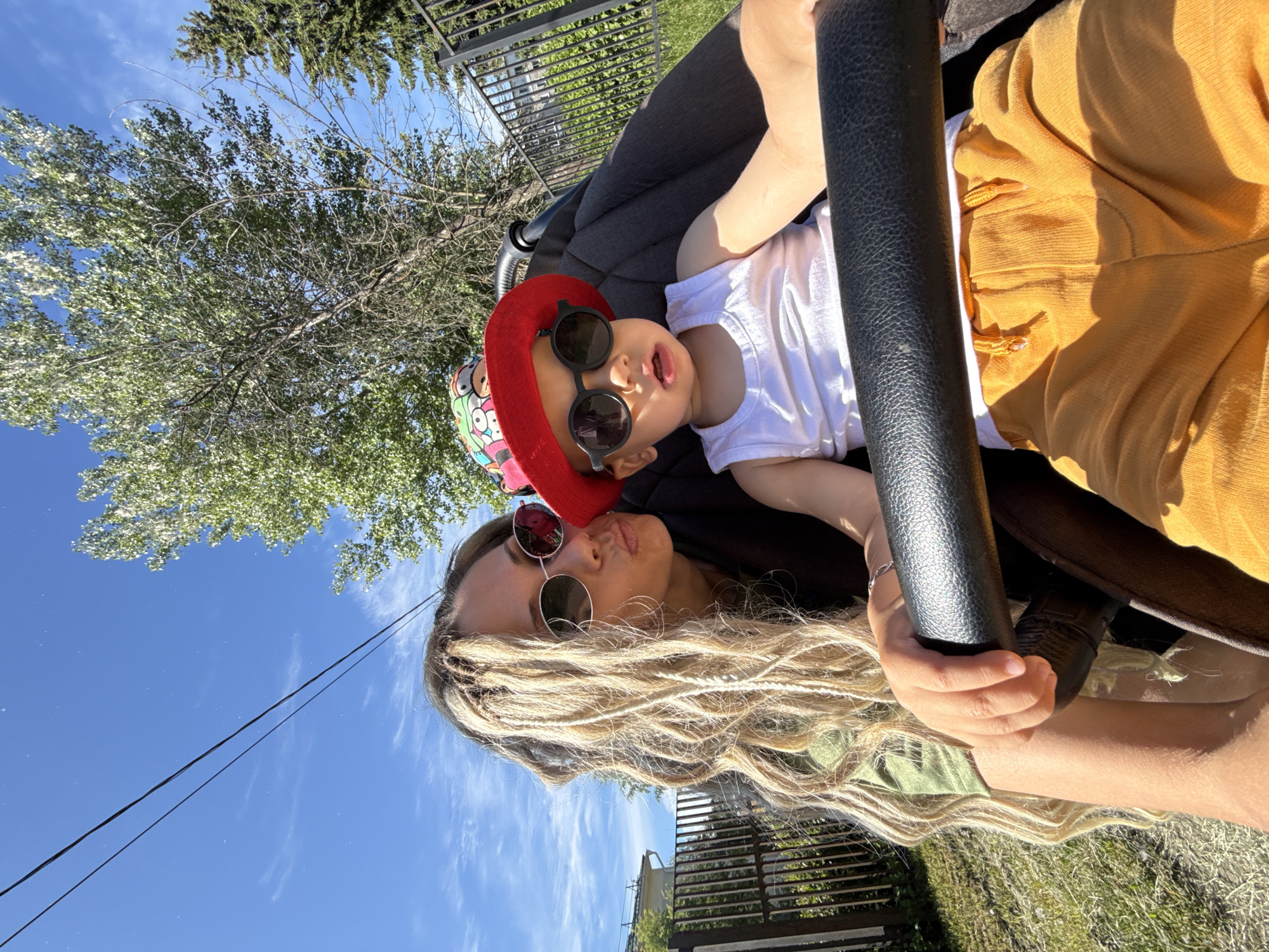 In the city of Rusanovo, Russia, a woman with blonde dreadlocks and round sunglasses puckers her lips towards a young child seated in a stroller. The child, wearing a red bucket hat and black sunglasses, is dressed in a white tank top and orange shorts. The stroller has a black handlebar. They are outdoors, with a bright blue sky and scattered clouds above. A tall tree with green leaves is visible in the background, along with a black metal fence and some buildings. The ground is covered in dry grass.