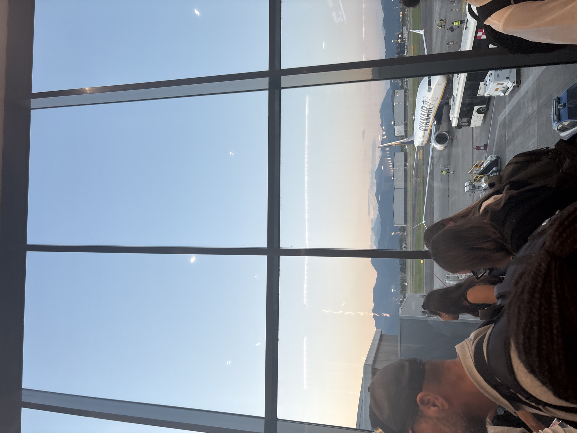 The image shows a view through a large window at an airport. In the foreground, several people are standing, facing away from the camera, suggesting they are waiting. One person in the center foreground is wearing a dark cap and a grey collared shirt, with a backpack visible. To their right, a woman with long dark hair is looking upwards.
Outside the window, on the tarmac, a Ryanair airplane is parked. A white bus is positioned near the airplane, and other airport vehicles and personnel are visible. In the background, mountains are silhouetted against a pale blue and orange sky, indicating either dawn or dusk. The airport appears to be Orio al Serio International Airport (also known as Il Caravaggio International Airport), located near Bergamo, Italy, given the presence of Ryanair, a major airline operating there, and the characteristic mountain backdrop often seen from this airport.