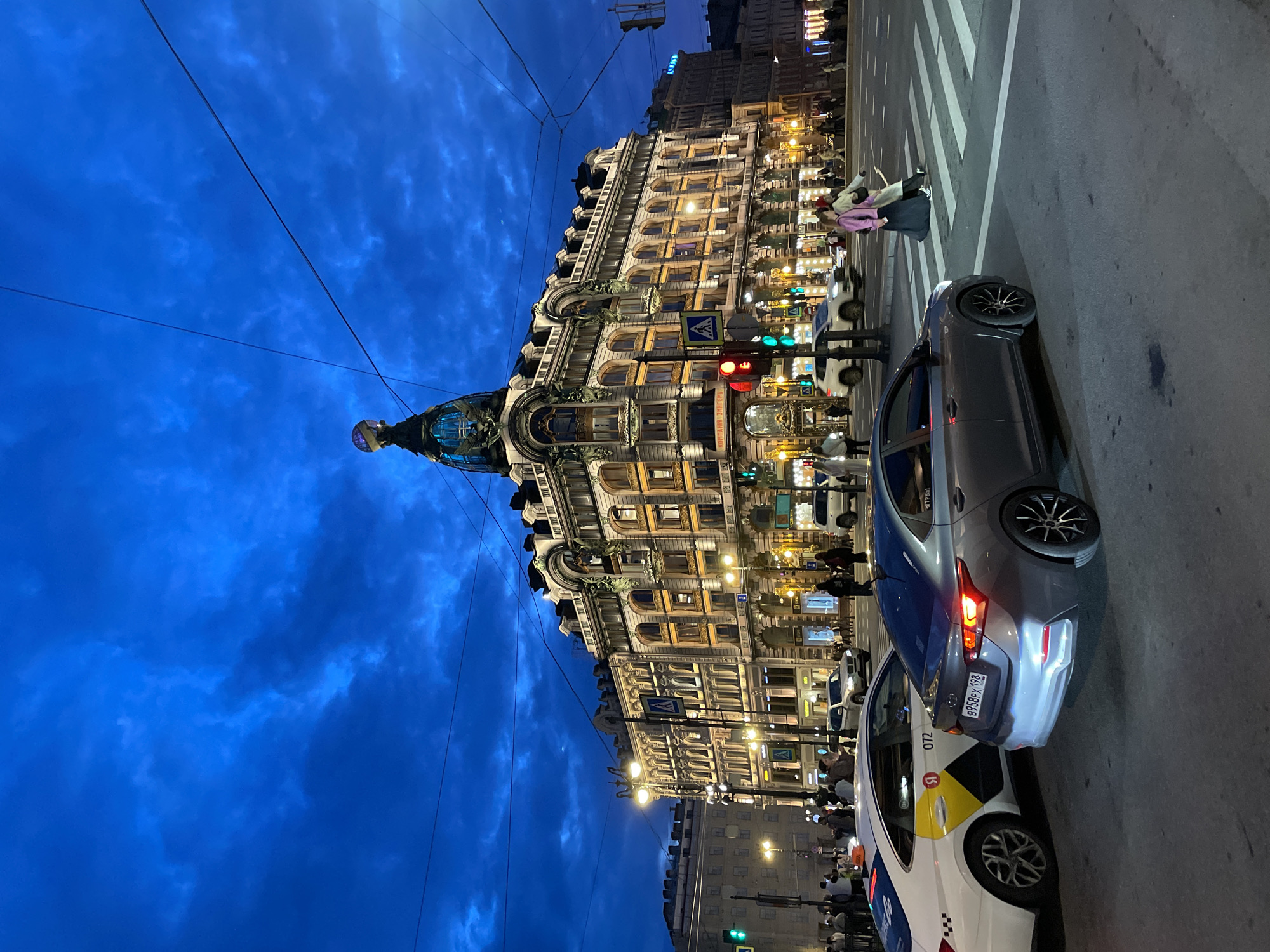 In the city of Unknown, Unknown, a view of a grand, ornate building with a distinctive domed structure at its peak is captured at dusk. The building is illuminated with warm, artificial lights, highlighting its architectural details. The sky above is a deep, dramatic blue, filled with textured clouds. In the foreground, a multi-lane intersection is active with traffic. Several cars are visible, including a white taxi with yellow and black markings on its side, and a dark gray sedan. Pedestrians are crossing the street on a marked crosswalk. Traffic lights are visible, with a red light illuminated on one of the signals. Overhead wires crisscross the sky, a common urban feature. The scene conveys a sense of a vibrant city at twilight.