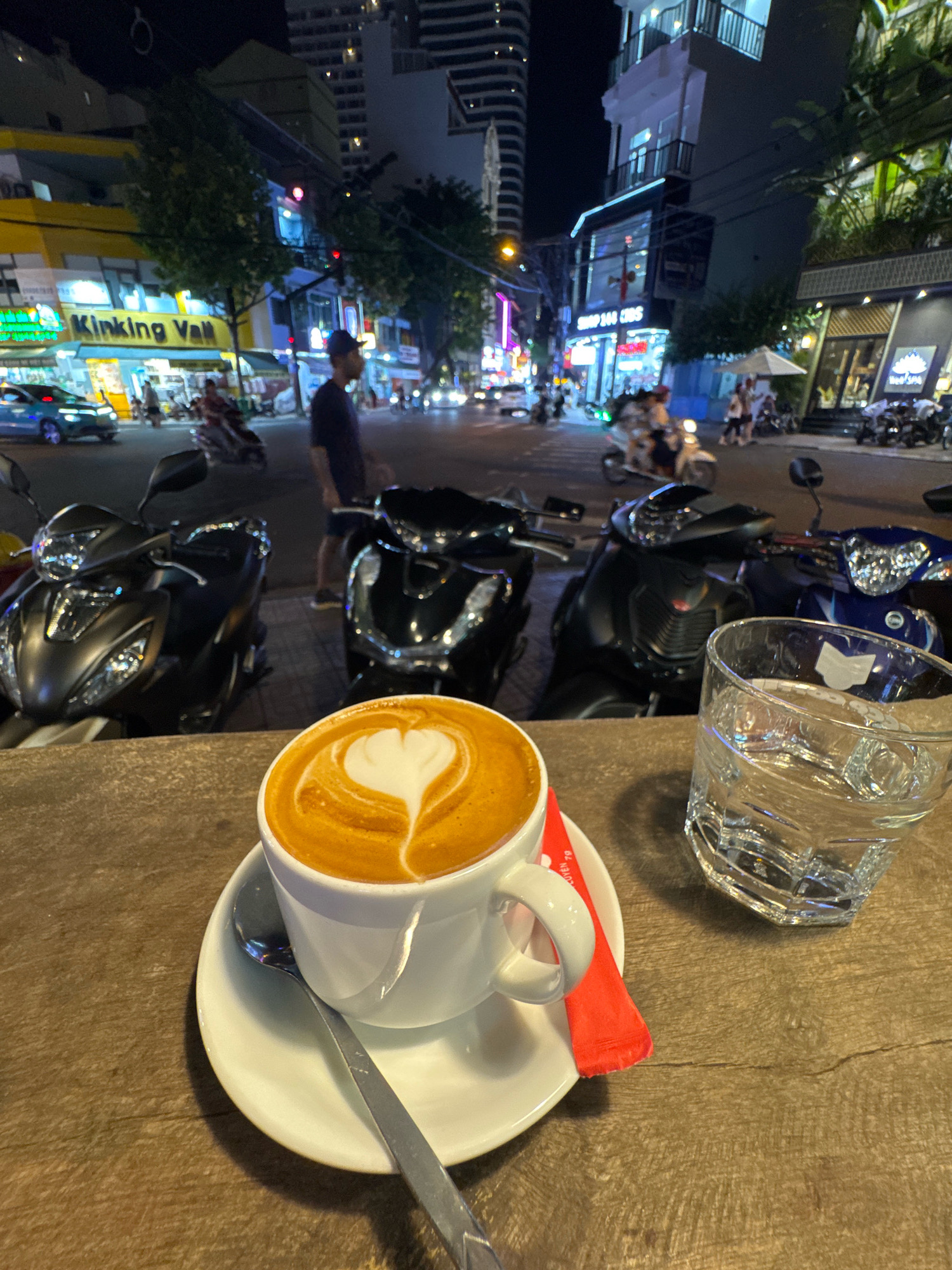 A close-up, eye-level outdoor shot features a white cup of latte art coffee with a heart shape, resting on a saucer with a spoon and a red sugar packet on a wooden table. To the right, a clear glass filled with water and ice sits on the table. In the background, a street scene unfolds at night in Nha Trang, Vietnam, with multiple motorbikes parked and moving, pedestrians, and illuminated buildings with various signs. The scene is likely captured on a busy street near the city center, possibly in the tourist district, given the mix of local and commercial establishments.