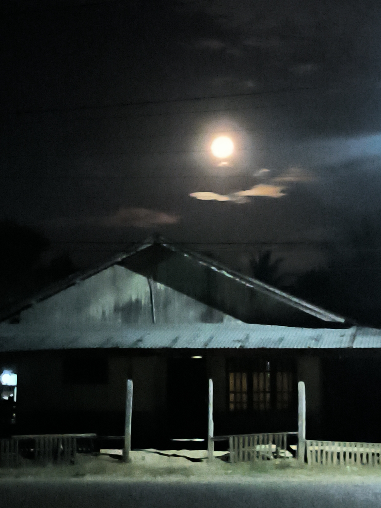 A low-angle, poorly lit, outdoor nighttime shot shows a single-story house with a gabled roof, located in Merbopuntong, Indonesia. A bright, full moon is visible through scattered clouds in the dark sky above the house. The house features a corrugated metal roof, a porch supported by four wooden posts, and a low picket fence. The windows of the house are dark, with only the outlines of vertical bars visible. A streetlight is visible in the far left, and sparse foliage and other objects are visible in the background. A road is visible at the bottom of the image.