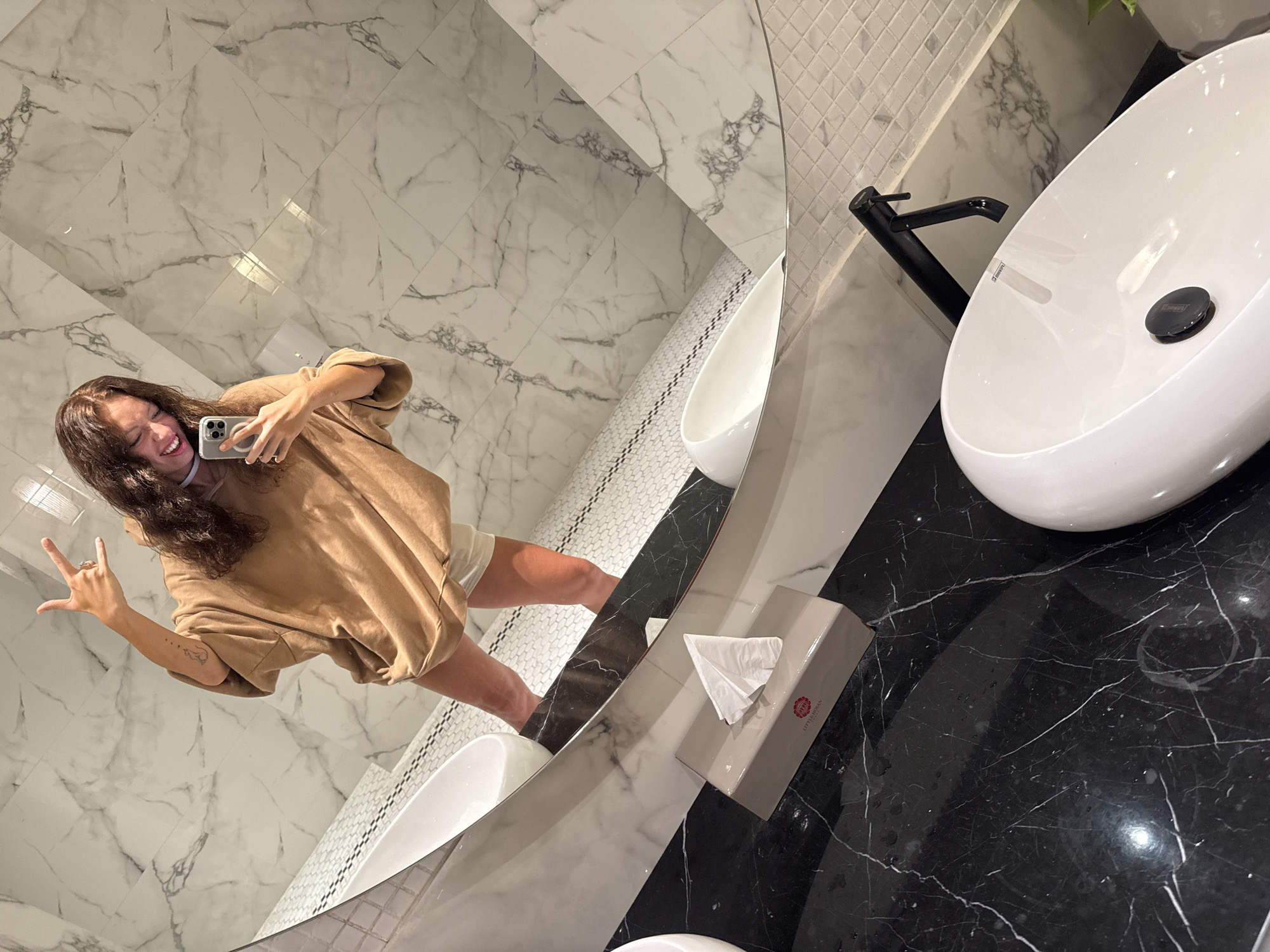 A mirrored selfie captures a woman in a modern bathroom. The bathroom features marble walls, a white ceramic urinal, and a black marble countertop with a white sink. A tissue box sits on the counter. The woman, wearing a tan shirt and light-colored shorts, holds a phone in her right hand and makes a "rock on" hand gesture with her left. Based on the logo on the tissue box, this photograph was taken at the Little Hoi An, located in Đà Nẵng, Vietnam.