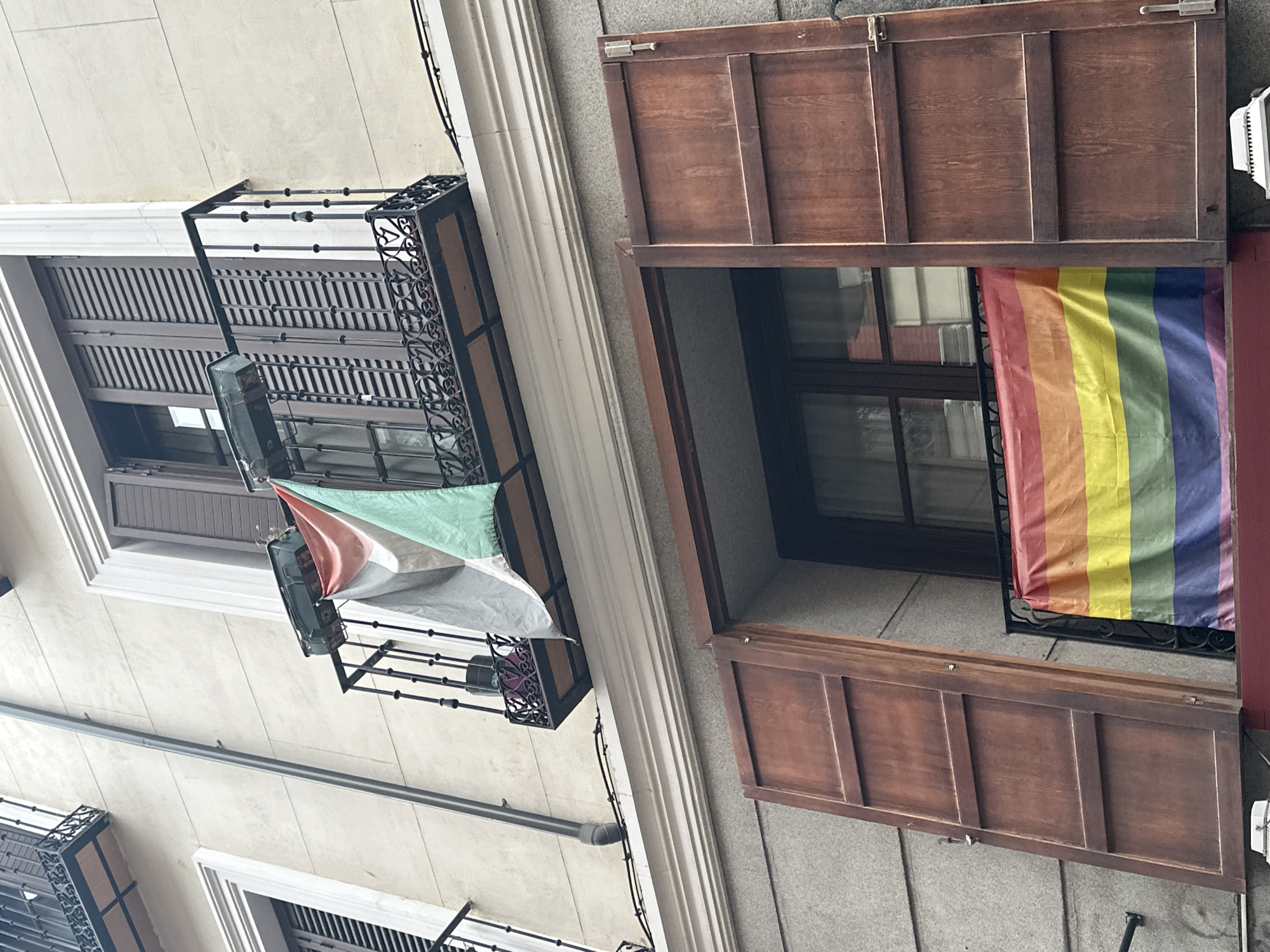 The image shows a building facade in Madrid, Spain. There are two windows with balconies. The upper balcony has a dark metal railing and a window with closed brown shutters. A flag with red, white, and green stripes hangs from the railing. The lower window has open brown shutters and a rainbow flag hanging from its railing. The building appears to be constructed of stone and stucco. The overall impression is of a residential building with decorative elements.
