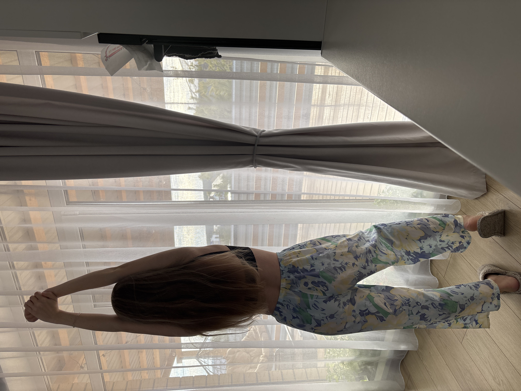 The photograph shows a person, likely female, stretching with arms extended overhead, back towards the camera, indoors, with a window behind them. The person has long brown hair and wears a black top and floral pants. They also wear fuzzy slippers. The window is covered by light-filtering white sheer curtains and heavier gray curtains tied back in the middle. Outside the window, trees are visible. The location is likely indoors, in a residence. The room has wooden floors and is well-lit. The city and country cannot be ascertained from the image.