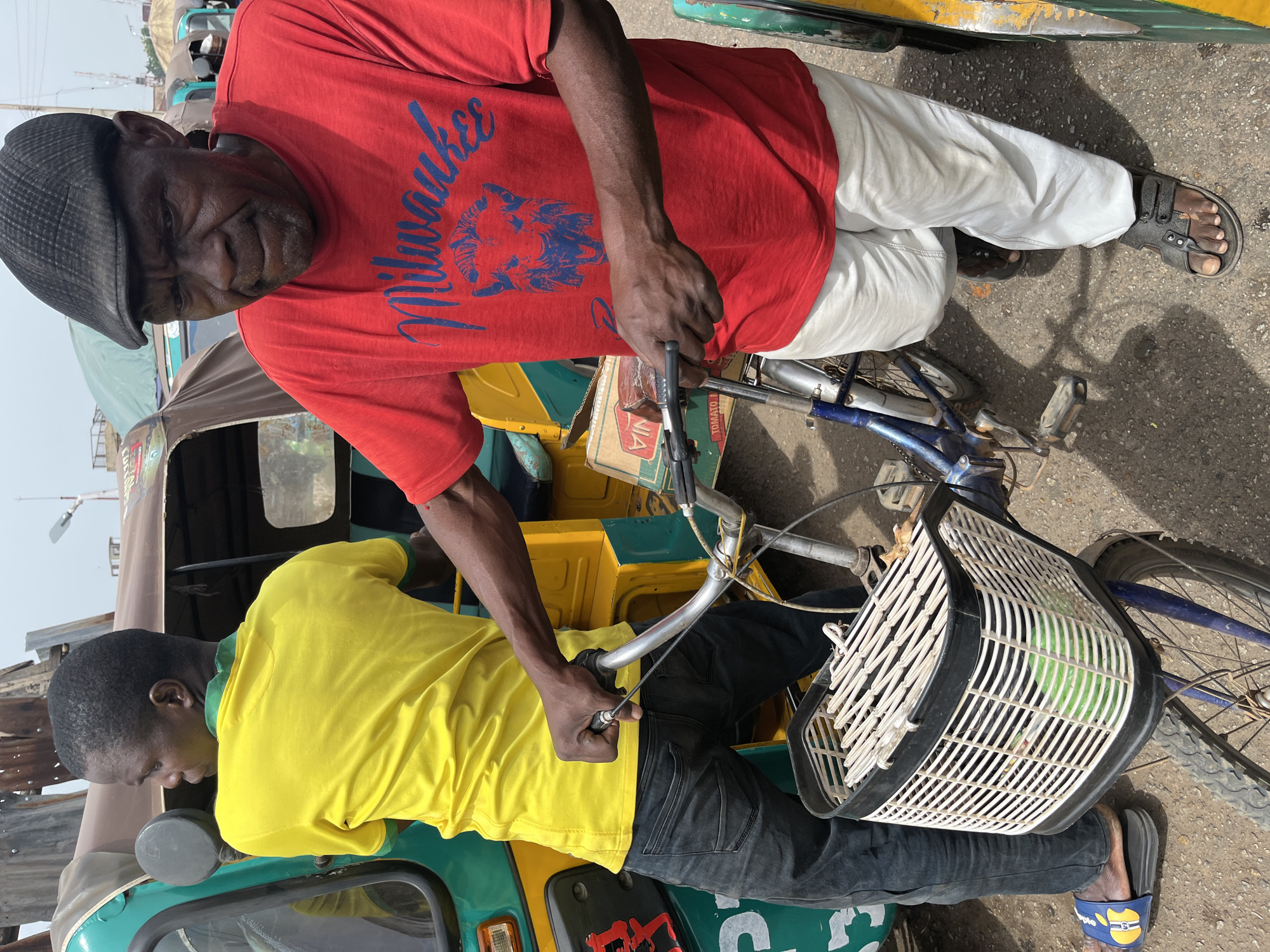 Two men are interacting with a bicycle equipped with a front basket. The man in the red "Milwaukee" T-shirt and white pants is standing behind the bicycle, holding the handlebars. The second man, wearing a yellow T-shirt and black pants, is standing in front of the bicycle, also holding the handlebars. The bicycle is positioned between the two men, and in front of a yellow and green auto rickshaw. The scene appears to be taking place on a street in Jos, Nigeria.