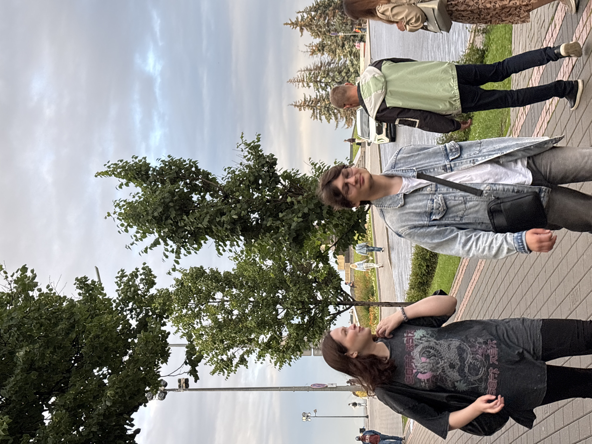 The image depicts an outdoor scene in Nizhny Novgorod, Russia, possibly near a waterfront or park. In the foreground, a young woman with long brown hair is walking from left to right, wearing a dark graphic t-shirt and black leggings. To her right, a young man with short brown hair and glasses is walking in the same direction, wearing a light blue denim jacket over a white t-shirt and grey pants, with a black crossbody bag. Behind them, a man in a green and black jacket is walking away from the camera, with a white car visible further in the background. In the distance, several people are walking on a paved pathway lined with greenery and trees. The sky is overcast.