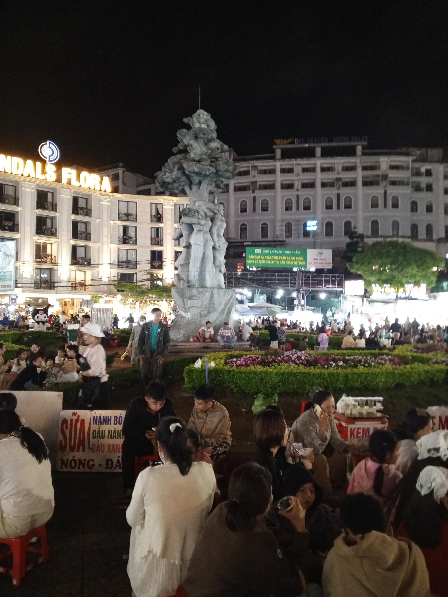 The image captures a nighttime scene in Da Lat, Vietnam, likely at a public square or market area. In the background, a multi-story white building with illuminated windows and a neon sign reading "NDALS FLORA" stands prominently. Adjacent to it, a large, grey stone sculpture resembling a tree with figures is centrally positioned. Further in the background, another building displays a sign that reads "DASAVICO HOTEL DALAT". The foreground is populated with numerous people, some seated on small red stools, others standing or walking. A row of colorful flowers and green bushes forms a landscaped barrier in the mid-ground. Several individuals appear to be interacting with each other, with some holding phones and others gathered in small groups. Two prominent signs are visible: one with Vietnamese text in red and white, and another with the word "KEM" (ice cream in Vietnamese) in red. The atmosphere suggests a lively evening gathering with food and social interaction.
