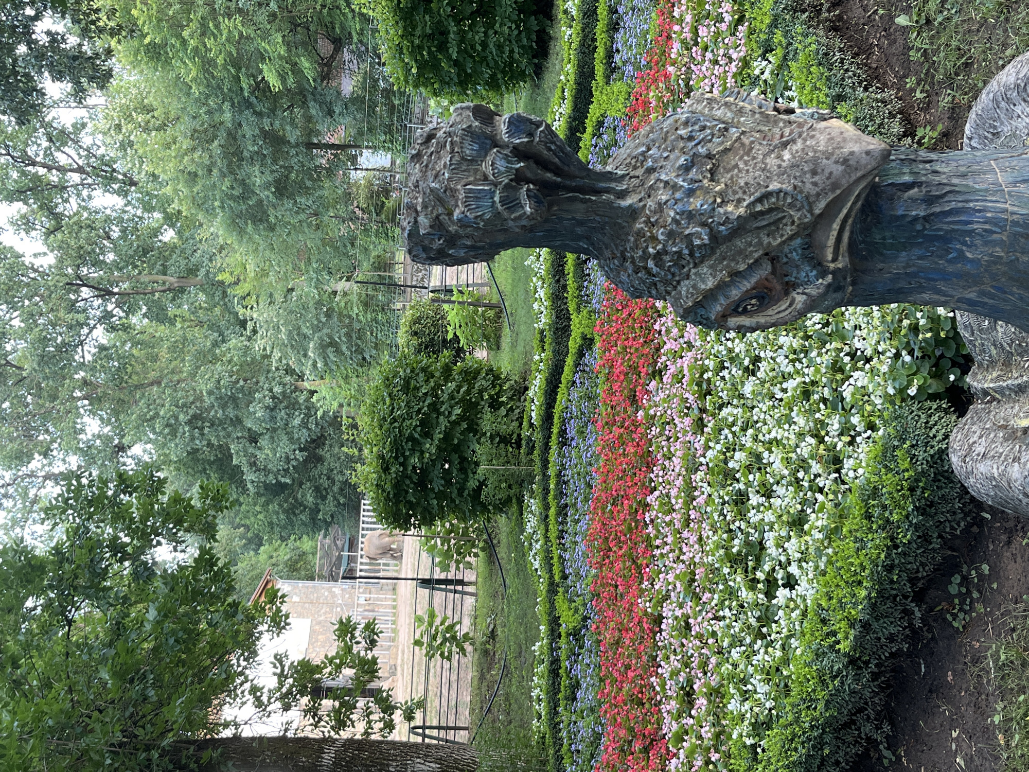 The image depicts an outdoor scene in the Nyíregyháza Animal Park, also known as Sóstó Zoo. In the foreground, a weathered, abstract sculpture of a bird's head, likely a peacock given the textured crown-like element, is prominently displayed. The sculpture is dark blue and grey, with visible texture suggesting it's made of stone or a similar material. Behind the sculpture, a meticulously maintained flower bed displays rows of vibrant flowers in shades of red, pink, white, and purple, bordered by neatly trimmed green hedges. In the middle ground, an elephant is visible in an enclosure, characterized by a fence and a light-colored building in the background. The background consists of lush green trees, indicating a natural or park-like setting. The overall atmosphere is one of a well-kept zoo or botanical garden.