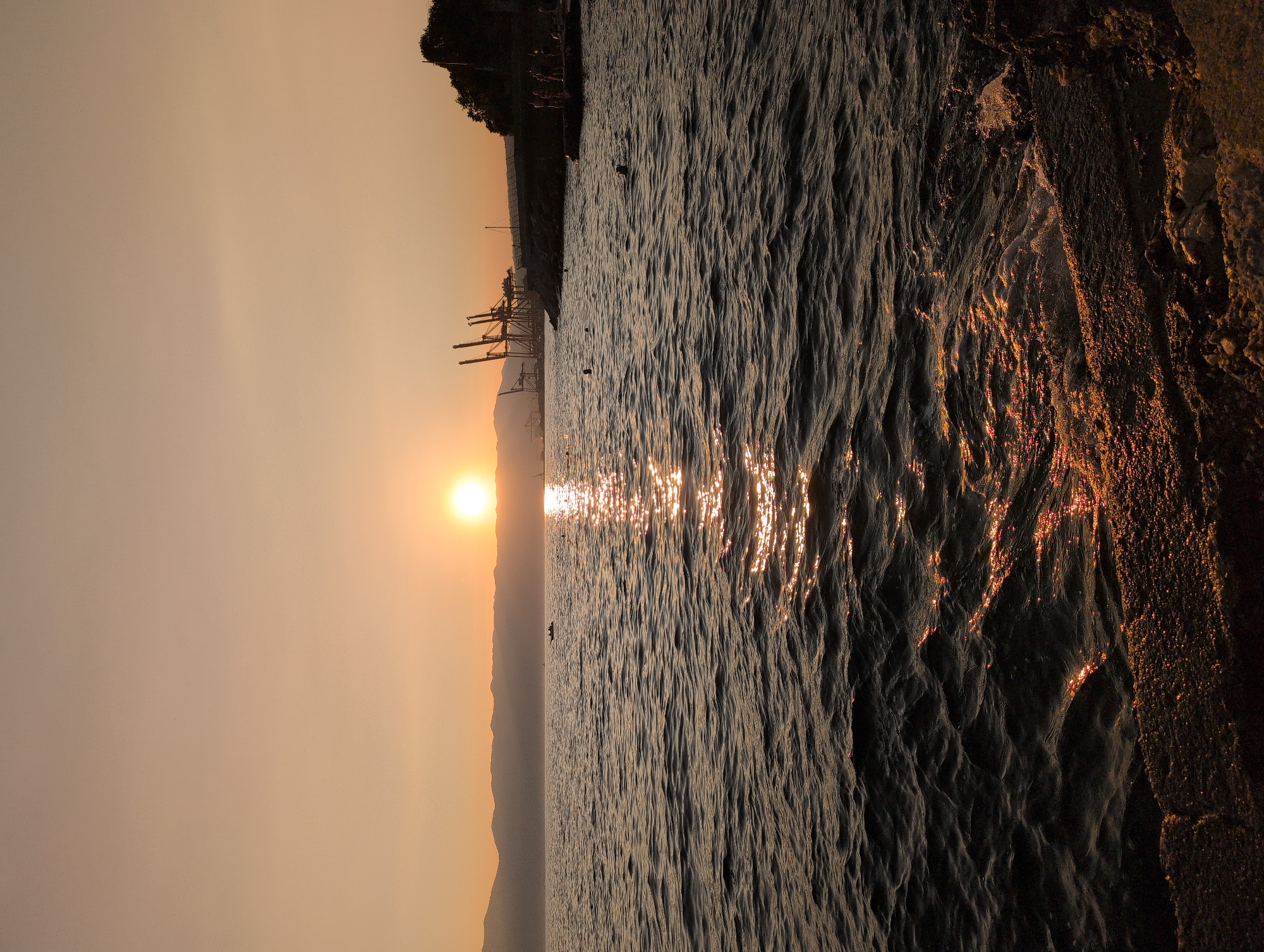 The image depicts a sunset over the Adriatic Sea in the city of Rijeka, Croatia. The sun is low on the horizon, casting a warm, golden glow across the sky and reflecting on the rippling water. In the distance, a chain of hills forms the coastline, and a few boats are visible on the sea.
On the right side of the image, a harbor area is visible, with several large cranes dominating the skyline. A tree-covered hill rises behind the industrial structures, and a rocky shoreline with a few people standing or swimming is present in the foreground. The water is dark, with the sunlight creating a shimmering path across its surface. The overall scene suggests a tranquil evening at a coastal city, with a mix of natural beauty and industrial activity.