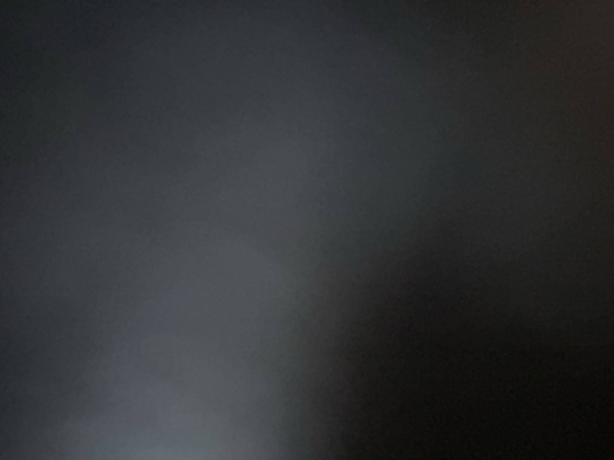 The media file is a photograph of a dark, monochromatic surface with subtle gradients of gray. There are no discernible subjects such as people, objects, or a clear scene. The lighting appears to be dim, with a gradient that transitions from a lighter gray on the left side to a darker gray and black on the right. This suggests a low-light environment or a surface that absorbs light. There are no visible location cues, text, or indications of time of day or weather. The overall impression is of an abstract, minimalist image with a focus on texture and light. Given the provided location context of Belgrade, Serbia, it is possible this depicts a wall, a piece of furniture, or another dark surface within an indoor or outdoor setting in the city, though no specific details confirm this. The lack of any defined features makes it impossible to determine the activity or event taking place.