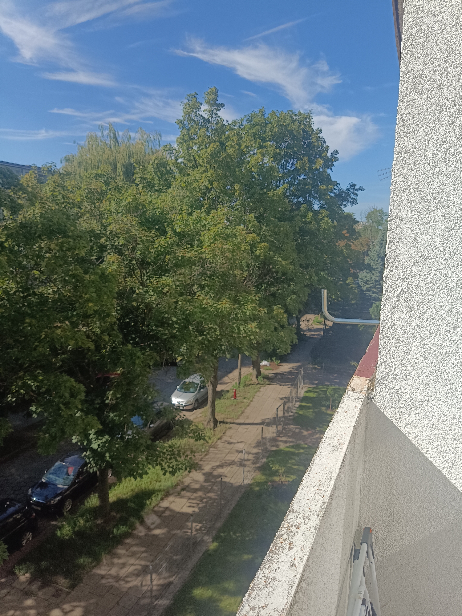 An outdoor, eye-level, medium shot captures a view from a balcony, looking down onto a residential street with trees and parked cars, under a blue sky with white clouds. The balcony railing is weathered and peeling. To the right, a textured white wall of a building is visible. A metal pipe extends horizontally from the building, curving upwards. Below, a paved sidewalk lines the street, bordered by a strip of grass. Several trees, with dense green foliage, cast shadows on the sidewalk and street. Two cars are parked on the side of the street: a dark sedan closest to the bottom left, and a silver hatchback further up the street. A red fire hydrant is visible near the silver car. The general urban environment suggests a residential area.