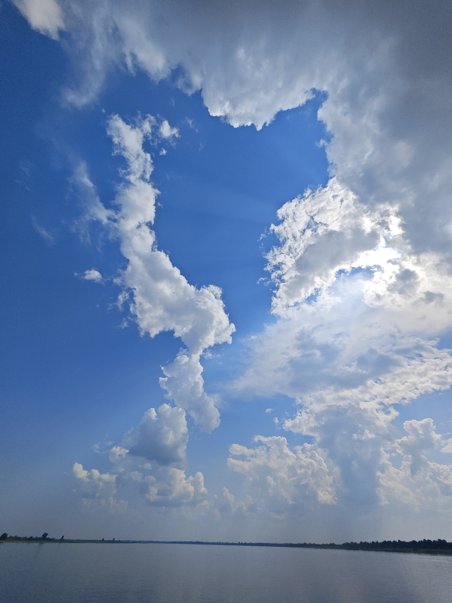 The image depicts a vast, calm body of water, likely a river or lake, stretching towards a distant horizon. The water is a deep blue-grey, reflecting the sky above. Along the far bank, a thin line of dark green vegetation, possibly trees or reeds, marks the edge of the land. The sky is a brilliant blue, dominated by a dramatic display of white and grey clouds. Sunlight pierces through a gap in the clouds, casting visible rays downwards, illuminating the scene with a soft, ethereal glow. The clouds are varied in texture and form, some dense and voluminous, others wispy and translucent. The overall impression is one of natural beauty and tranquility. There are no people or man-made structures visible in the image, with the exception of a subtle, dark line on the horizon that might represent distant land features. The scene is consistent with a natural landscape in the city of Erkalovo, Russia, given the presence of large bodies of water and a temperate climate that supports such cloud formations.