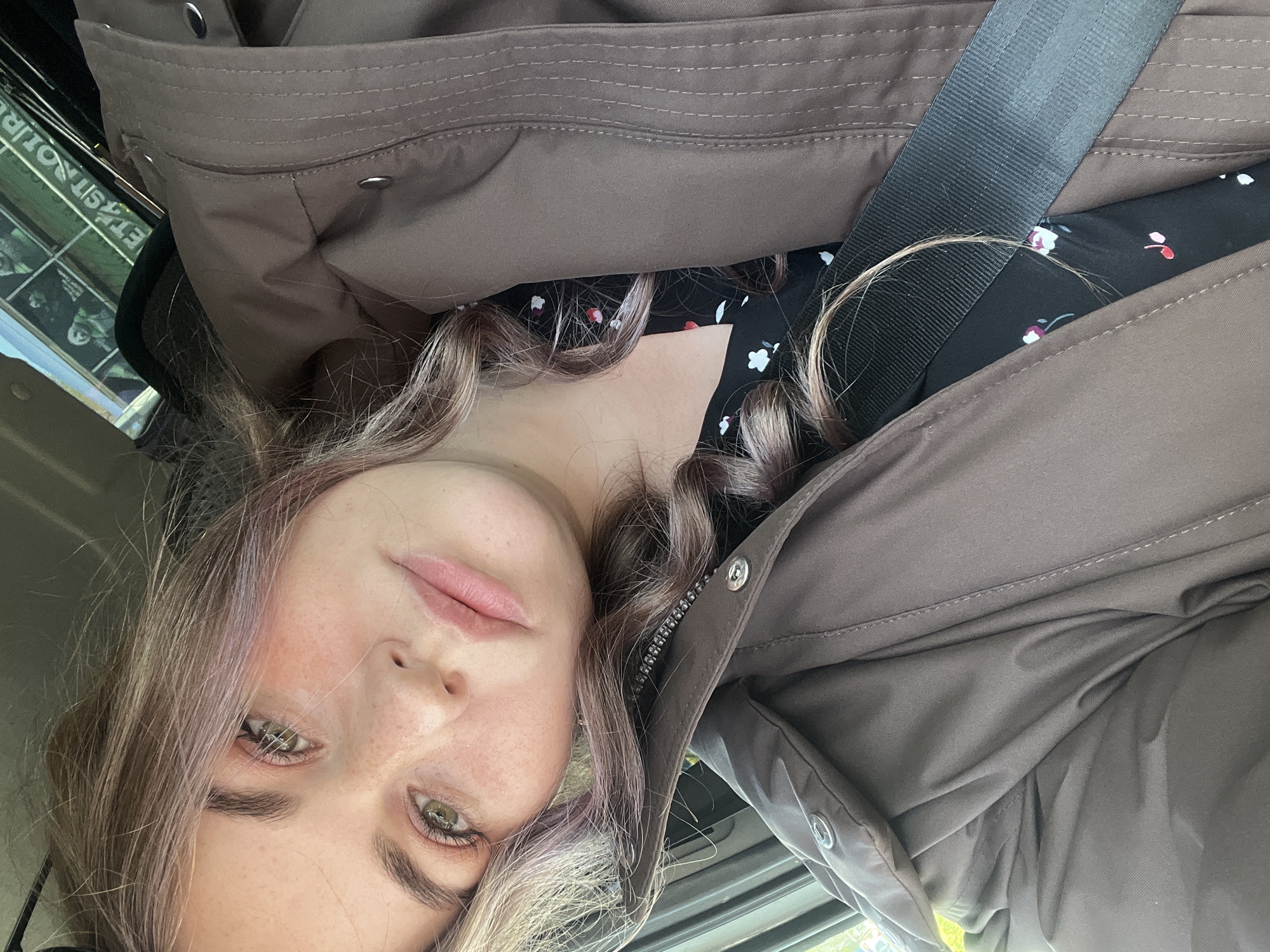 The image shows a selfie taken in what appears to be the inside of a car. A young woman with light brown hair, fair skin, and green eyes is looking at the camera. She is wearing a black dress with a small floral print, a brown jacket, and a seatbelt is diagonally across her. The background shows a window with a reflective surface, and part of some posters in what can be identified as the city of Chaikovsky, Russia. The lighting is natural.