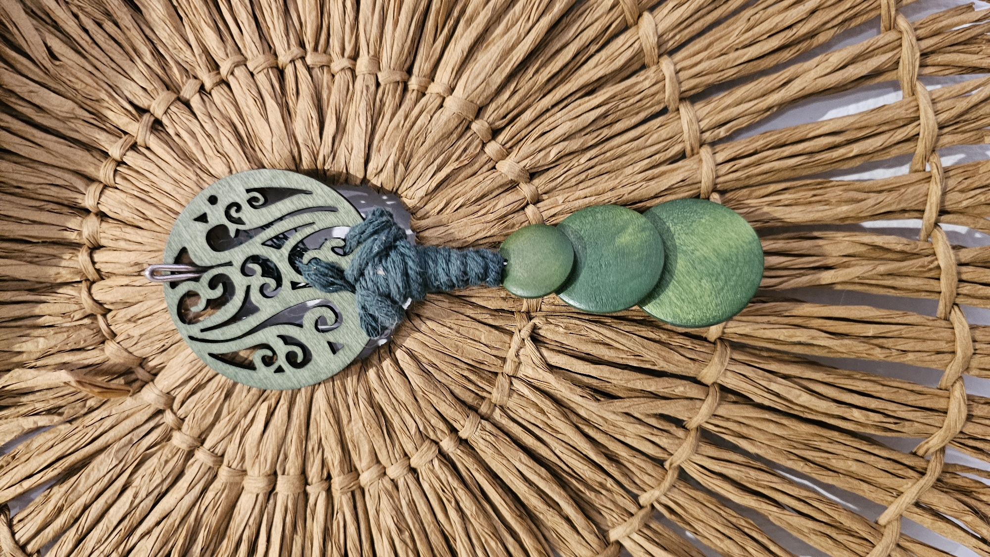 The image shows a close-up of a handcrafted item, likely a wall hanging or decorative piece. The background is composed of radiating strands of a light brown, textured material, possibly raffia or straw, tied at intervals with thinner strands of the same material. This creates a fan-like or sunburst pattern.
Centered in the image is a circular, light green wooden ornament with intricate cut-out designs. A silver-colored loop is attached to the top of this ornament, suggesting it's meant to be hung. Below the wooden circle, a thick, dark green cord is tied in a knot and then descends, wrapping around a series of three stacked, flat, circular wooden beads. These beads are also a shade of green, with visible wood grain. The beads decrease in size from top to bottom, with the largest at the bottom.
The image is taken from a straight-on perspective, with a shallow depth of field that blurs the edges of the woven background slightly. The lighting appears to be natural and even, highlighting the textures of the materials. There are no people or discernible actions taking place in the image; it focuses solely on the object and its immediate surroundings. This decorative item is positioned against a white surface, which is partially visible at the bottom of the frame.
The prompt requests information about the location in Baan Chaweng, Thailand. However, there are no visual cues in the provided image or crops that indicate this specific location. The image solely depicts the decorative object itself and its immediate context, which is a white background. Therefore, no details about Baan Chaweng, Thailand can be derived from the visual information presented.