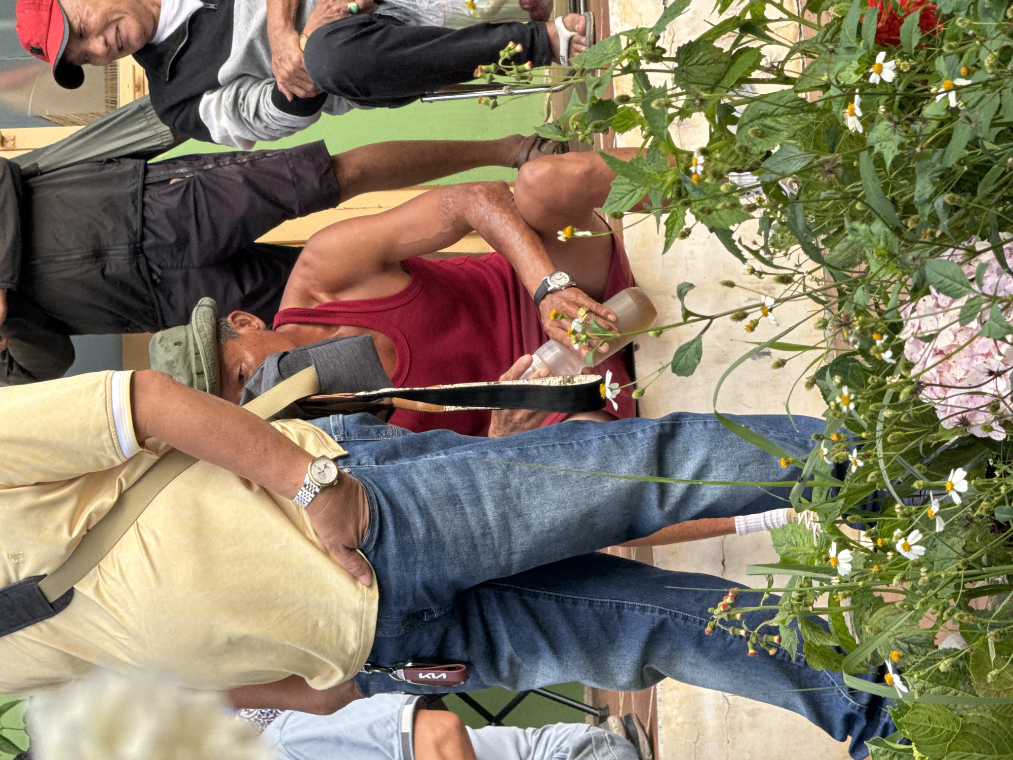 The image depicts a group of men in an outdoor setting, possibly a street or public space, in Da Lat, Vietnam. In the foreground, a man wearing a yellow polo shirt and blue jeans stands with his hand in his pocket, a watch on his wrist, and a key chain attached to his belt loop. Beside him, another man in a red tank top and a green hat sits down, holding a clear bottle of liquid. Further back, a man in a black jacket and dark shorts is partially visible, and a man in a red baseball cap and a grey and black jacket sits on a ledge, looking to the side. The background includes a green wall and a lighter-colored structure. Green foliage with small white flowers is prominent in the lower portion of the image, suggesting a natural or cultivated environment within the urban landscape of Da Lat. The overall impression is a casual gathering of individuals outdoors.