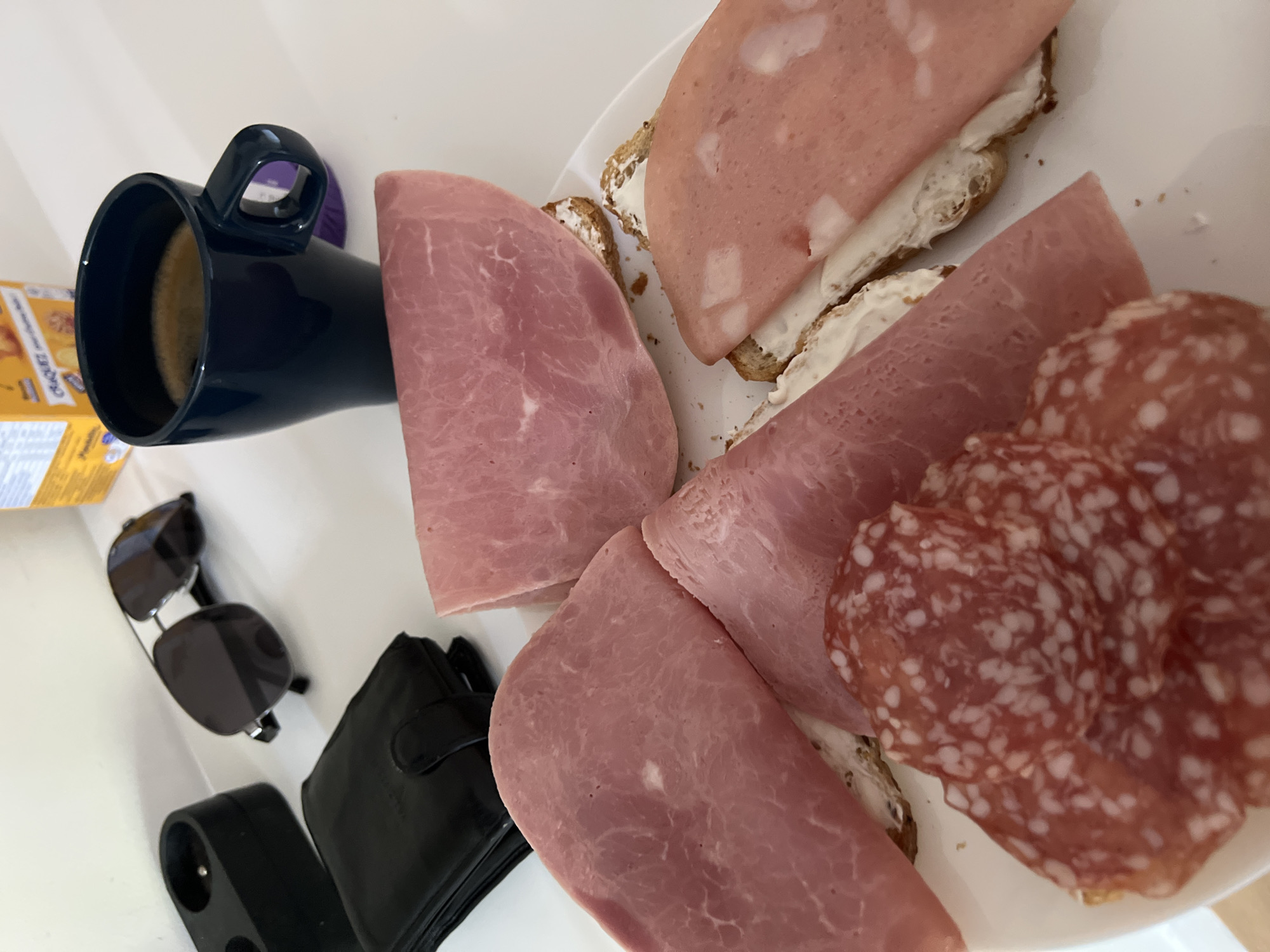 The image displays an overhead view of various items arranged on a clean white surface, likely a table. The central focus is a casual meal setup, primarily featuring a plate laden with deli meats and open-faced sandwiches.
On a white plate, several slices of pink deli meat, possibly ham or mortadella, are arranged, some folded and others topping small sandwiches. These sandwiches are made with brown bread, spread with a white creamy substance that resembles cream cheese. Additionally, there are multiple slices of red salami, distinctively dotted with white specks. Small crumbs are visible on the plate, suggesting it has been used.
To the left of the plate, a dark blue mug with a handle holds a light brown beverage, likely coffee or tea. Further to the left, a yellow and orange cardboard box is partially visible, with text fragments that appear to say "CHEES" and "SNACKS," indicating a product like crackers or cheese snacks.
Below these items, a pair of black sunglasses with dark lenses and silver accents is folded. Adjacent to the sunglasses is a closed black wallet. At the bottom left, a black electrical outlet extension or adapter with two sockets is present.
The scene suggests a daytime meal or snack, possibly breakfast, in an indoor setting such as a home or office. No people are visible in the image, and no specific location cues are discernible to confirm Vitry-sur-Seine, France.