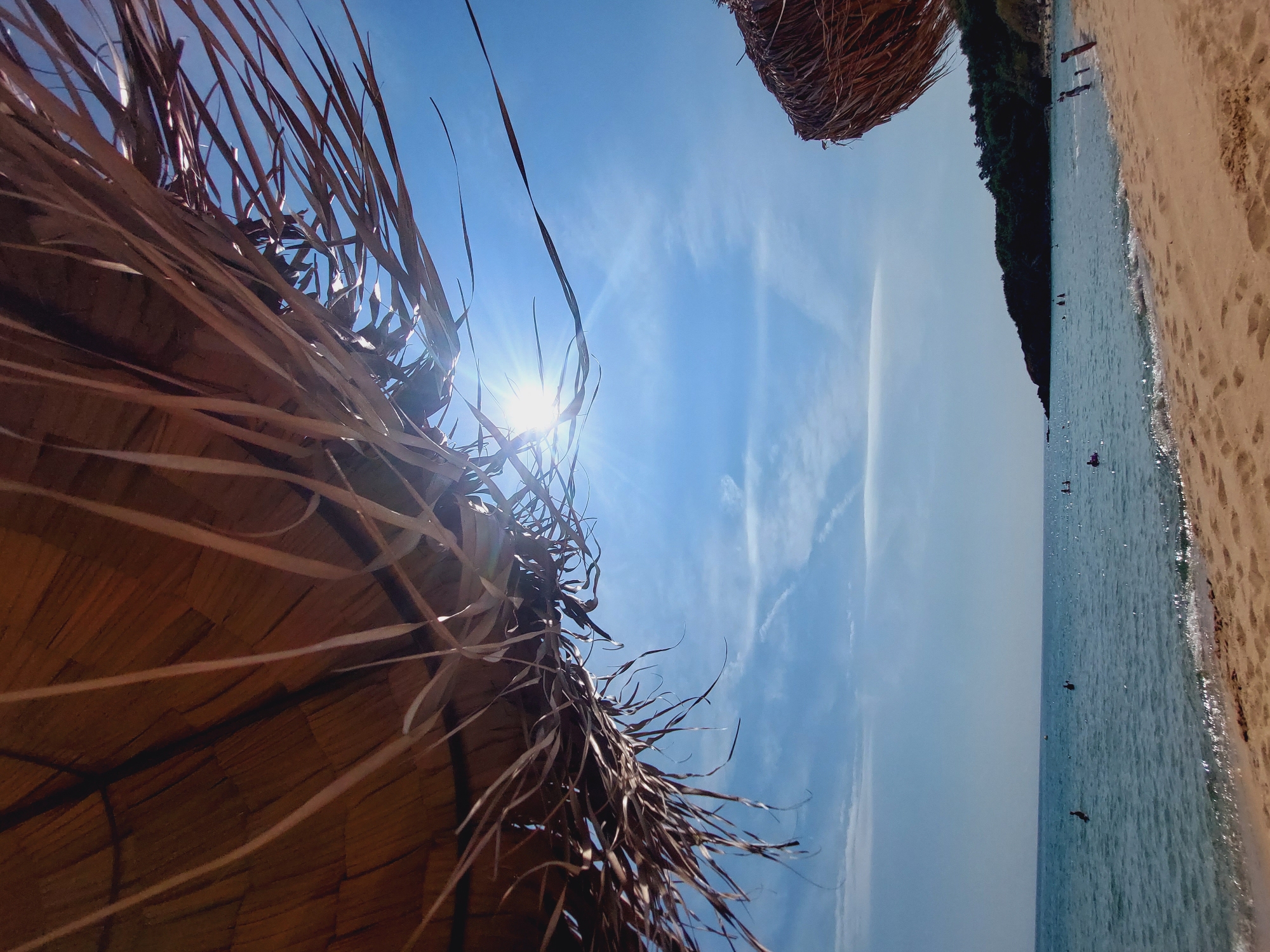 This is a ground-level, low-angle, wide shot of a sunny beach scene, possibly in Sozopol, Bulgaria, taken from beneath a thatched umbrella. The foreground is dominated by the rough texture of the umbrella's straw material, which obscures a significant portion of the upper frame and casts a partial shadow. Sunlight pierces through the straw, creating lens flare and highlighting the translucent nature of the dried plant matter. The middle ground features a sandy beach with gentle waves lapping at the shore. Several people are visible in the shallow water, some swimming or wading, while others appear to be further out. The water is a clear, inviting blue, reflecting the bright sky. In the background, a lush, tree-covered headland juts out into the sea, forming a natural boundary to the cove. The sky is a vibrant blue with scattered white clouds, suggesting fair weather. Another thatched umbrella is partially visible on the right side of the frame, indicating a beach with amenities. The overall impression is one of a peaceful, idyllic summer day at the coast.