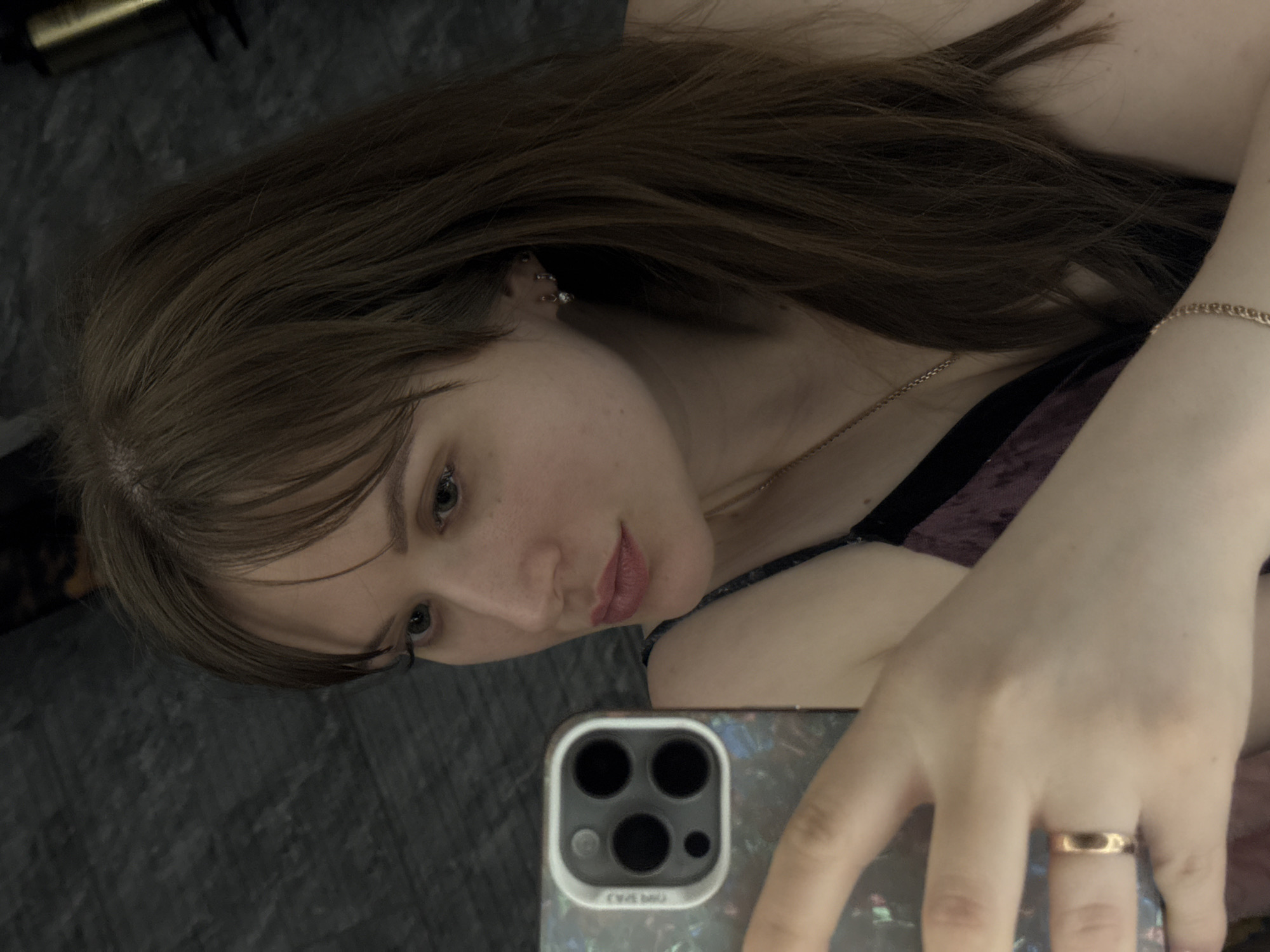 The image shows a woman with long brown hair and light skin taking a selfie. She is wearing a gold ring and a gold bracelet. She has a gold necklace. The phone she is using has three camera lenses. The background appears to be a dark grey surface. There is no evidence to suggest the image was taken in the city of Chaikovsky, Russia.