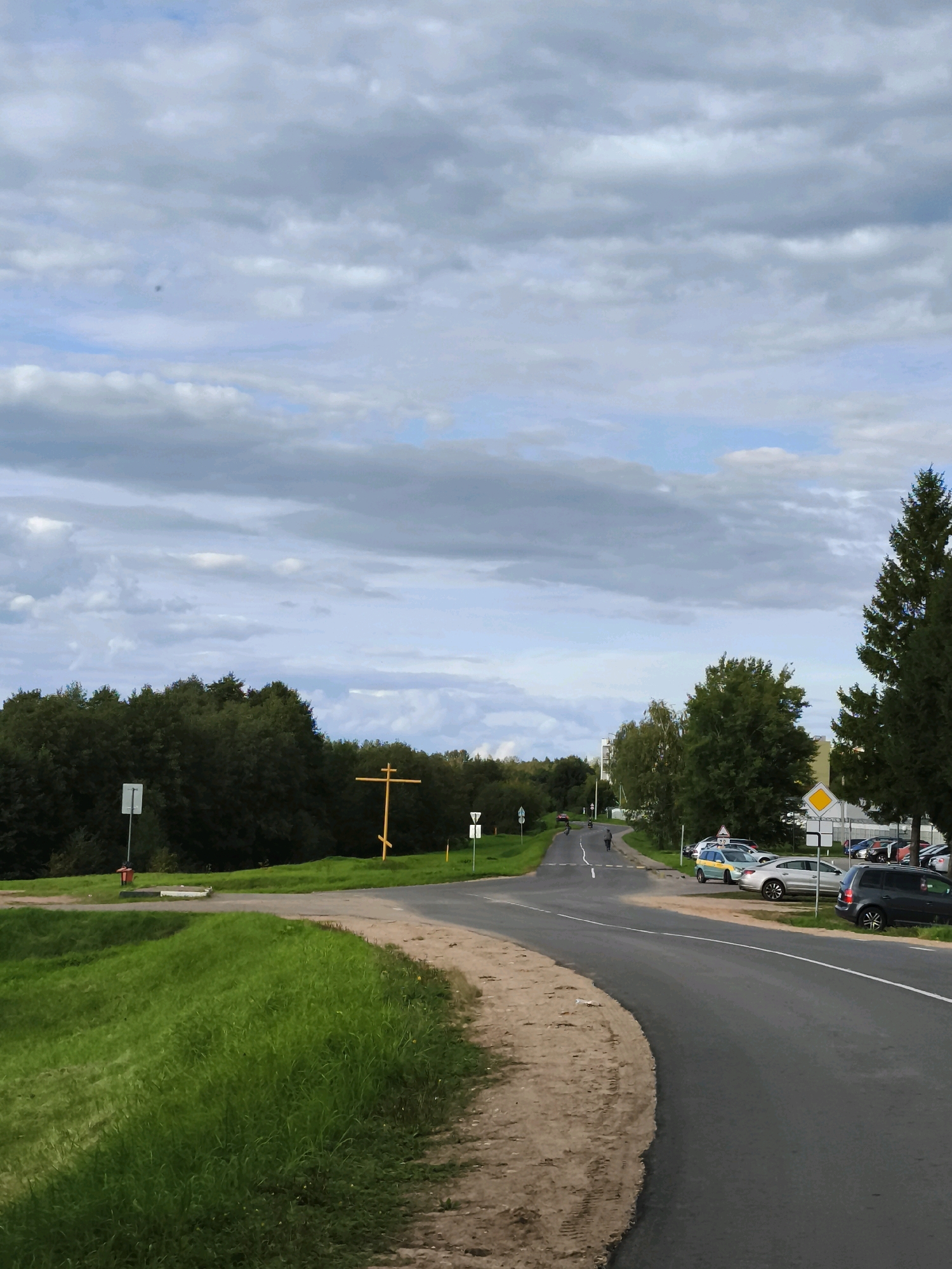 The image depicts a rural road scene in the vicinity of Горскі сельскі Савет, Belarus. The road curves to the right, with a grassy embankment on the left and a line of parked cars and trees on the right. In the distance, several figures can be seen walking or cycling along the road. A prominent wooden cross stands on the left side of the road, near a traffic sign and a fire hydrant. The sky is overcast with scattered clouds.