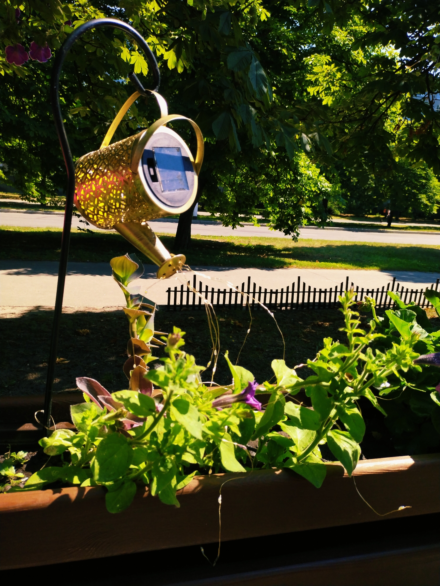 A decorative watering can solar light hangs from a curved metal stand in the foreground, set against a backdrop of lush green trees and a road. The watering can is gold and features a perforated, intricate pattern that allows light to shine through. A solar panel is visible on its top. Delicate string lights hang from the spout of the watering can, cascading down towards a planter box filled with vibrant green plants and a few purple flowers. A low, decorative black fence runs horizontally in the middle ground, separating the planter area from a grassy strip and a paved road beyond. In the far background, a few people can be seen walking along the road, suggesting a park or public space. The bright sunlight and dappled shadows indicate it is a sunny day, likely in the late morning or afternoon. The overall scene evokes a peaceful, garden-like atmosphere in an urban or suburban setting. The location is Horishni Plavni, Ukraine. There is no visible text in the image.