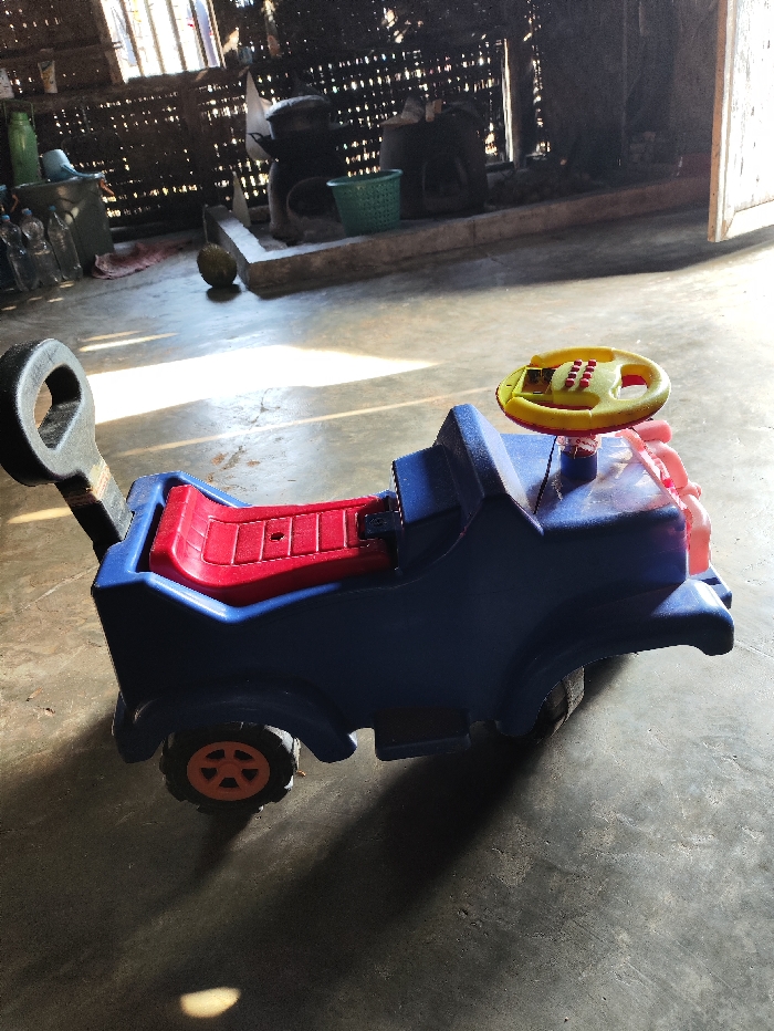 A bright indoor scene features a blue plastic ride-on toy car as the primary subject, positioned slightly off-center in the foreground. The toy car has a red seat, a yellow steering wheel with red buttons, and orange-colored wheels. A black handle or push bar extends from the rear of the vehicle.
The setting appears to be a simple, rustic interior, likely a room within a dwelling. The floor is made of concrete or a similar hard, grey material, marked with shadows cast by objects and sunlight. Strong, natural light streams in from the left side of the frame, creating prominent diagonal shadows across the floor, suggesting it is either morning or late afternoon on a sunny day.
In the background, against a wall made of woven material (possibly bamboo or rattan slats), various household items are visible. These include several clear plastic water bottles, a green plastic bucket or basket, and what appear to be traditional cooking implements or large pots resting on a raised platform. A single, round green fruit, possibly a coconut, lies on the floor towards the back. The overall impression is of a modest living space. No people are present, and no specific activity is taking place; the scene is static. There is no visible text in the image. The environment aligns with the provided location context of Selagai Lingga, Indonesia, suggesting a rural or traditional domestic setting.