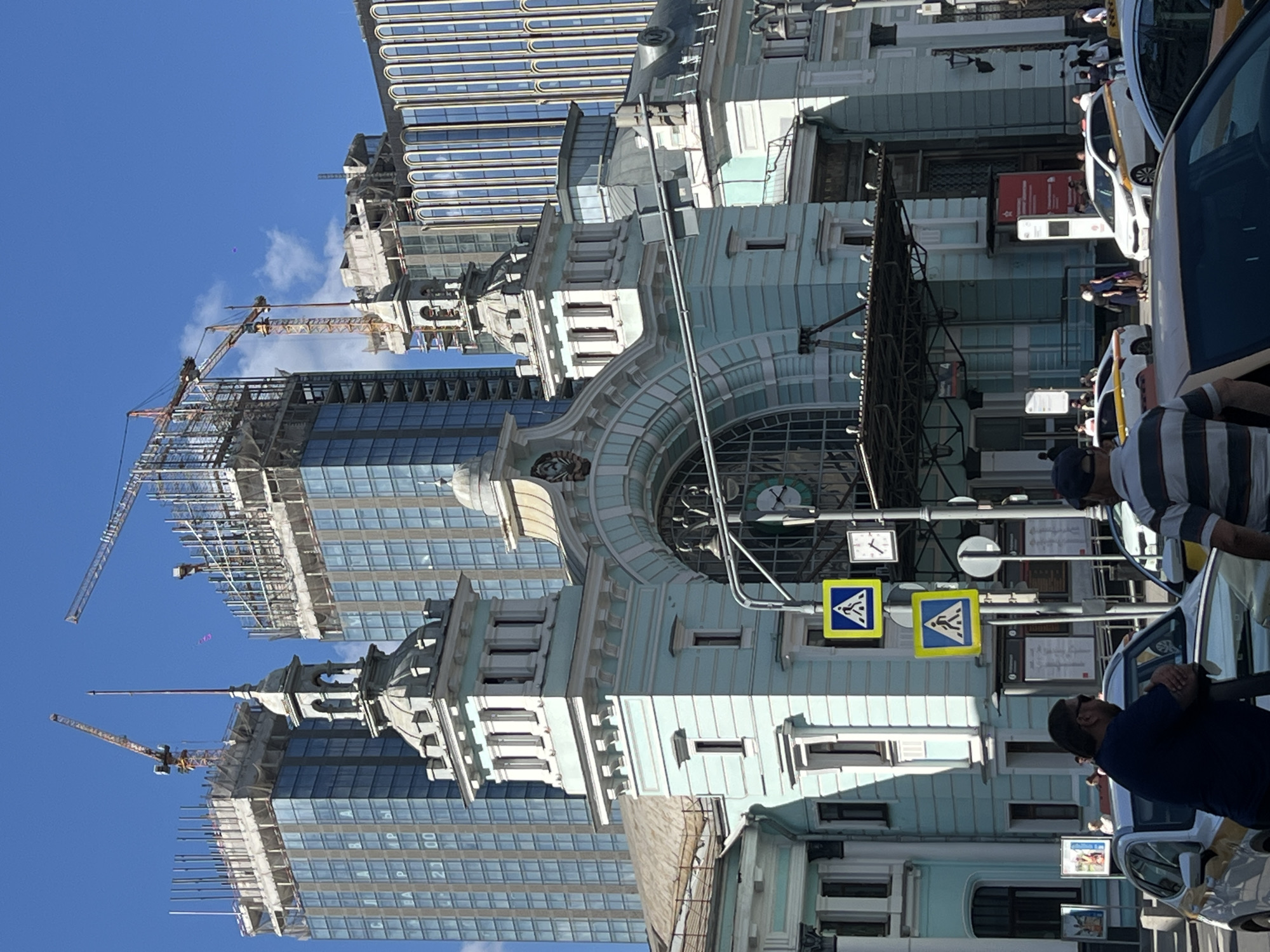 The image depicts a street scene in Moscow, Russia, where modern construction contrasts with historical architecture. A modern glass skyscraper under construction is visible in the background, with two prominent cranes extending from its incomplete structure. Juxtaposed against this is an older, ornate building, possibly dating back to the Tsarist era or pre-Soviet period, featuring detailed architectural elements and a large clock face. Street-level elements include pedestrian crossing signs and a cluster of individuals, some standing near vehicles, suggesting a bustling street environment. The sky is clear and blue, indicating fair weather conditions.