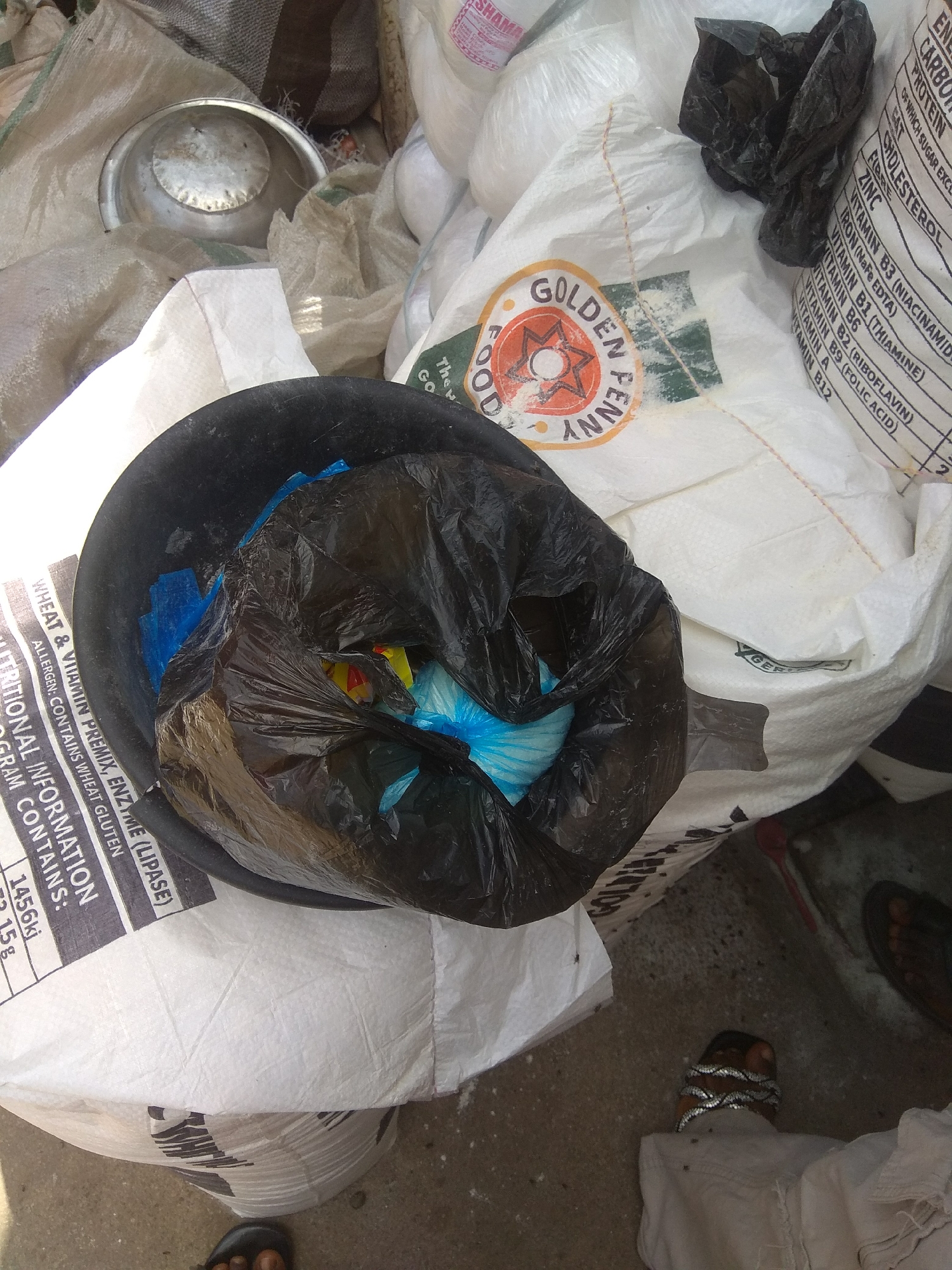 In Jalingo, Nigeria, a ground-level outdoor scene depicts a collection of discarded items and some personal belongings. The foreground is dominated by a black plastic trash bag filled with other smaller colored plastic bags, including blue and yellow. This trash bag is placed within a black circular container, possibly a bucket or bin, which is partially obscured by the trash bag itself.
Behind the trash, large white woven sacks are piled up. One of these sacks has visible nutritional information printed on it in black text. Text visible includes "WHEAT & VITAMIN PREMIX, ENZYME (LIPASE)", "ALLERGEN: CONTAINS WHEAT GLUTEN", and "PROGRAM CONTAINS", followed by numerical data. Another sack features a circular logo with text that appears to read "GOLDEN FOOD". A crumpled black plastic item, possibly another piece of trash or packaging, rests on top of the white sacks.
To the left, a metallic bowl is visible amongst the sacks. The ground is a rough, grey surface, likely concrete or compacted earth, with scattered debris. In the lower right corner, the feet of at least two individuals are present. One person's bare feet with sandals are visible, and another person's feet are adorned with what appears to be a coiled silver-colored bracelet around the ankle and toes, and they are wearing light-colored trousers. The lighting suggests it is daytime.