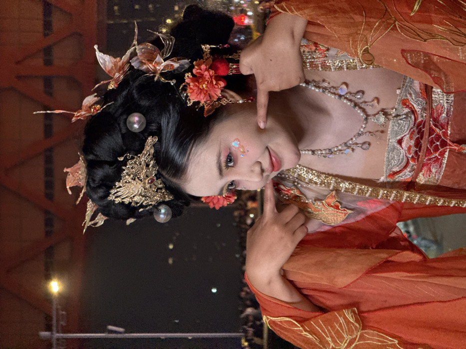 A young East Asian female is captured from the chest up, positioned slightly to the right of the frame's center, against a dark, illuminated urban night background. She wears an elaborate traditional Chinese-style costume predominantly in shades of orange and red, featuring a gold and white embroidered top with a prominent red floral motif. Her black hair is styled in a voluminous updo, adorned with intricate golden headpieces, including a large central ornament, smaller golden butterfly-like pieces, two iridescent spherical beads, and two red floral hairpins with dangling red elements. Her makeup includes bright pink eyeshadow, shimmering glitter on her upper cheeks, and light pink lipstick. She displays a slight smile, and her index fingers are pressed into her cheeks, creating artificial dimples. Around her neck is a multi-strand pearl necklace with iridescent and clear teardrop-shaped pendants and a central silver-toned charm. The sleeves of her costume are made of flowing, sheer orange fabric with golden embroidered patterns. The background consists of blurred yellow lights and indistinct figures, indicating a crowd. Above the crowd, a prominent architectural structure, possibly a bridge or large building, is illuminated with a strong orange-red light, featuring visible triangular structural patterns. This scene, particularly the illuminated bridge-like structure and city lights, is located in Yuzhong District, China, consistent with the nightscapes and architectural features of Chongqing.
