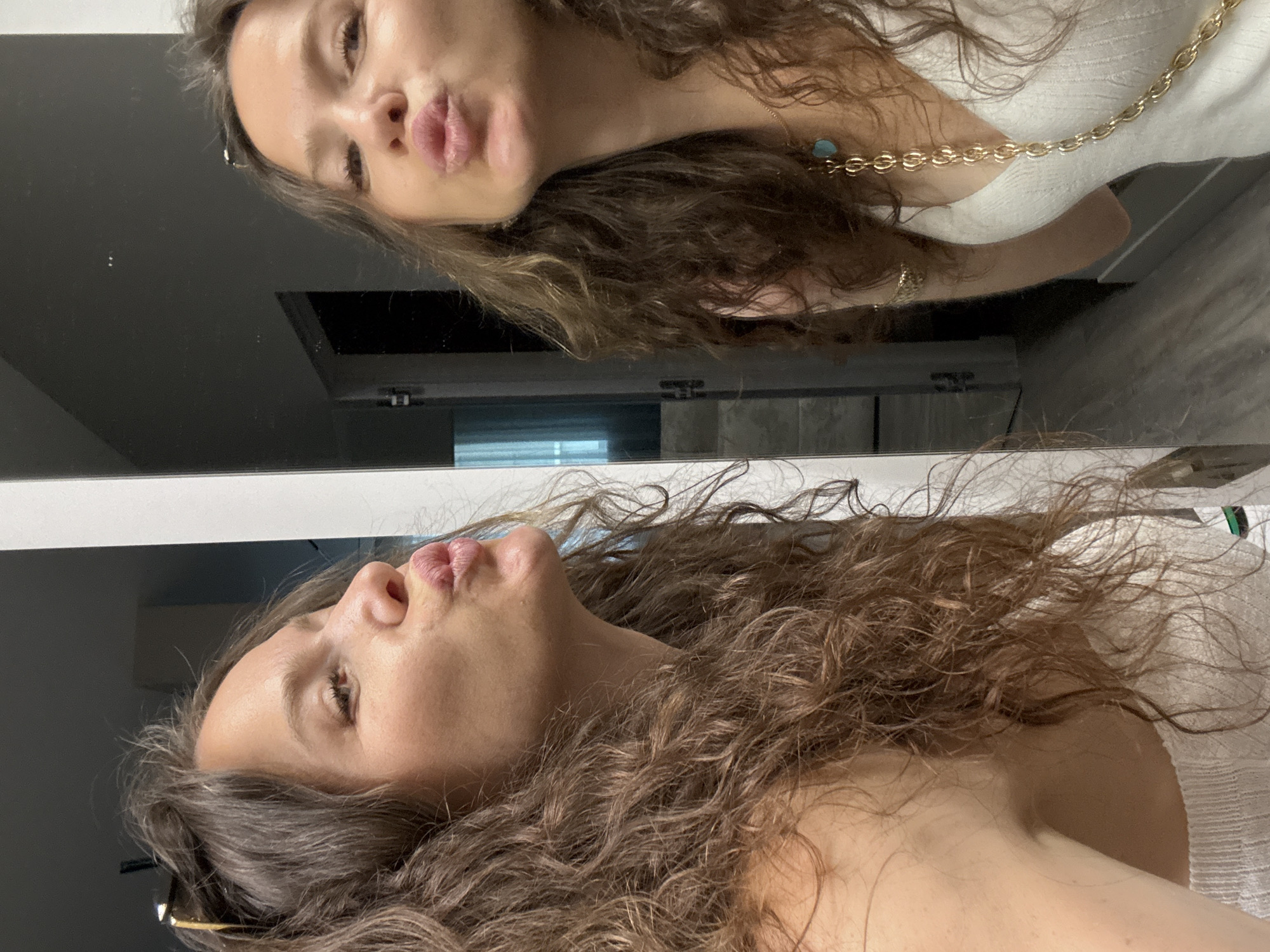In an indoor setting, a woman with long, curly brown hair is captured from the chest up in a selfie taken in front of a mirror. Her head is tilted to her left, and she is puckering her lips, looking towards the camera. She is wearing a white ribbed top and a gold chain necklace with a turquoise pendant. The mirror reflects her face and upper body, as well as a blurred background that includes a doorway, a staircase, and a tiled floor. The lighting is soft, casting a gentle glow on her skin.