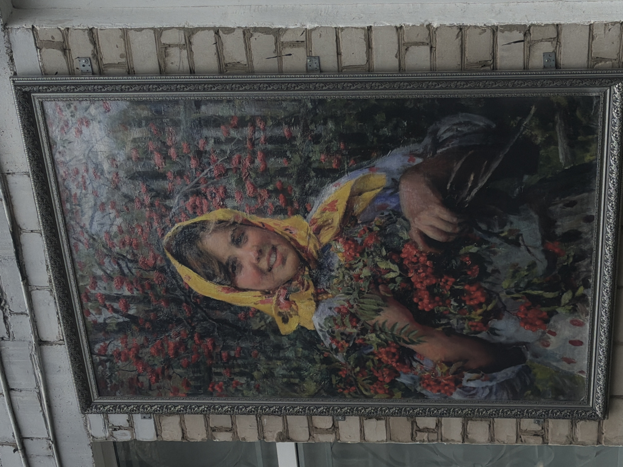 The image shows a framed oil painting hanging on a white brick wall, likely indoors. The painting depicts a young girl with a yellow headscarf surrounded by branches with red berries, potentially rowan berries. The background is a mix of green and red, suggesting foliage and more berries. The girl is smiling and appears to be holding or interacting with the branches. The frame is ornate and silver in color. The context of Chaikovsky, Russia, suggests the painting could be a local artwork or representation of rural life.