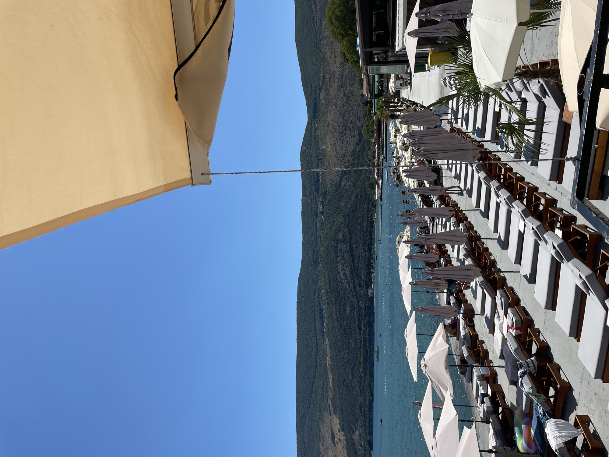 The image depicts a sunny day at a beach club in Igalo, Montenegro. Rows of empty, modern lounge chairs with gray cushions are arranged on a concrete promenade. Many of the chairs are shaded by large, light-colored umbrellas. The sea is visible in the background, with a few boats and swimmers. Beyond the sea, a lush, green, mountainous landscape stretches across the horizon. The upper right corner of the image is dominated by a portion of a beige awning. The scene appears to be set for a relaxed day at the beach, with most of the lounging areas unoccupied.