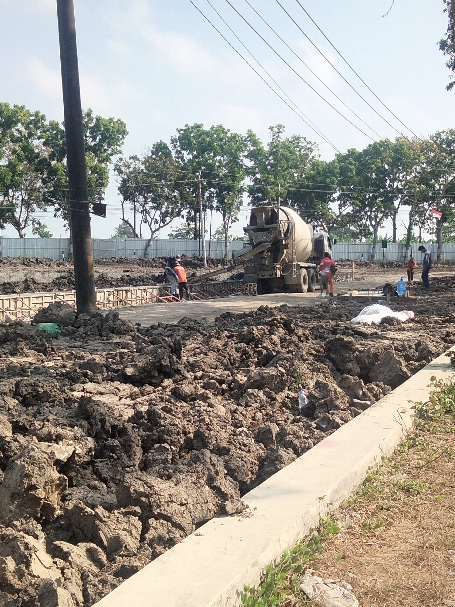 This is a medium-distance, eye-level shot of an outdoor construction site in Sikampuh, Indonesia. The foreground is dominated by a large pile of dark, muddy earth, with a lighter concrete structure running along the bottom right edge of the frame. In the mid-ground, a concrete mixer truck is visible, positioned on a paved area. Several construction workers are present. One worker, wearing an orange shirt and dark pants, is standing near the truck. Another worker in an orange shirt and dark pants is visible further to the right. A third worker, in a blue shirt and dark pants, is standing near a blue water cooler. A fourth worker, in a red shirt and orange pants, is near the truck. In the background, a line of trees forms a backdrop against a bright sky with scattered clouds. A tall, dark utility pole stands to the left of the frame. A light-colored metal fence runs along the edge of the property in the far background. There is also a small Indonesian flag visible on a pole in the far right background. The overall impression is of an active construction project.
