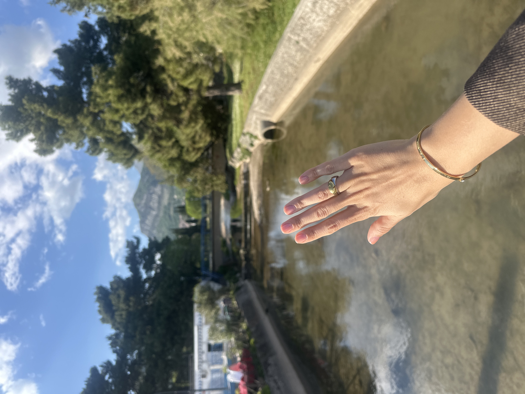 The image, oriented vertically, captures a bright outdoor scene featuring a person's right arm and hand extended into the foreground, reaching over a body of water. The hand is adorned with a gold-toned ring on the ring finger, and a slender gold-toned bracelet with a small jewel graces the wrist. The arm is covered by a textured, dark brown or grey sleeve.
The body of water, likely a canal or a narrow river, appears calm and reflects the sky, showing hues of blue and white clouds. A concrete bank with a visible drain pipe borders the water in the immediate foreground. Across the water, lush green trees and foliage line the banks. Further in the mid-distance, architectural elements are visible, including a prominent white building with red details and a distinct blue bridge.
In the background, majestic mountains rise under a bright blue sky dotted with scattered white clouds. The clear skies and strong lighting indicate it is daytime, likely midday or afternoon. The serene setting suggests a tranquil environment. Based on the provided context, the scene is located in Budva, Montenegro, highlighting its natural beauty and waterside features. No visible text is present in the image.