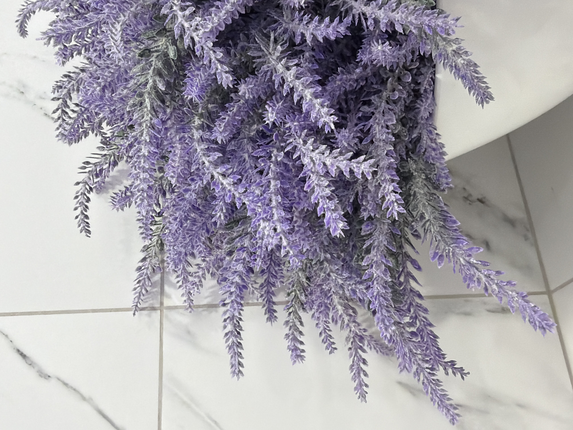 The image shows a close-up view of cascading artificial lavender flowers. The flowers are a vibrant shade of purple with white tips, giving them a frosted appearance. They are arranged in a dense cluster, with some sprigs hanging down. The background consists of white tiled walls, with faint gray lines suggesting grout. One of the tiles has a subtle marble-like pattern. The lighting appears to be artificial, possibly from an overhead source, creating soft shadows on the tiles. There are no people or animals visible. The scene is static, with no activity taking place. The overall impression is one of decorative floral arrangement, likely for indoor ambiance. The location context provided is Kazan, Russia, but there are no specific visual cues in the image to confirm this. There is no visible text in the image.