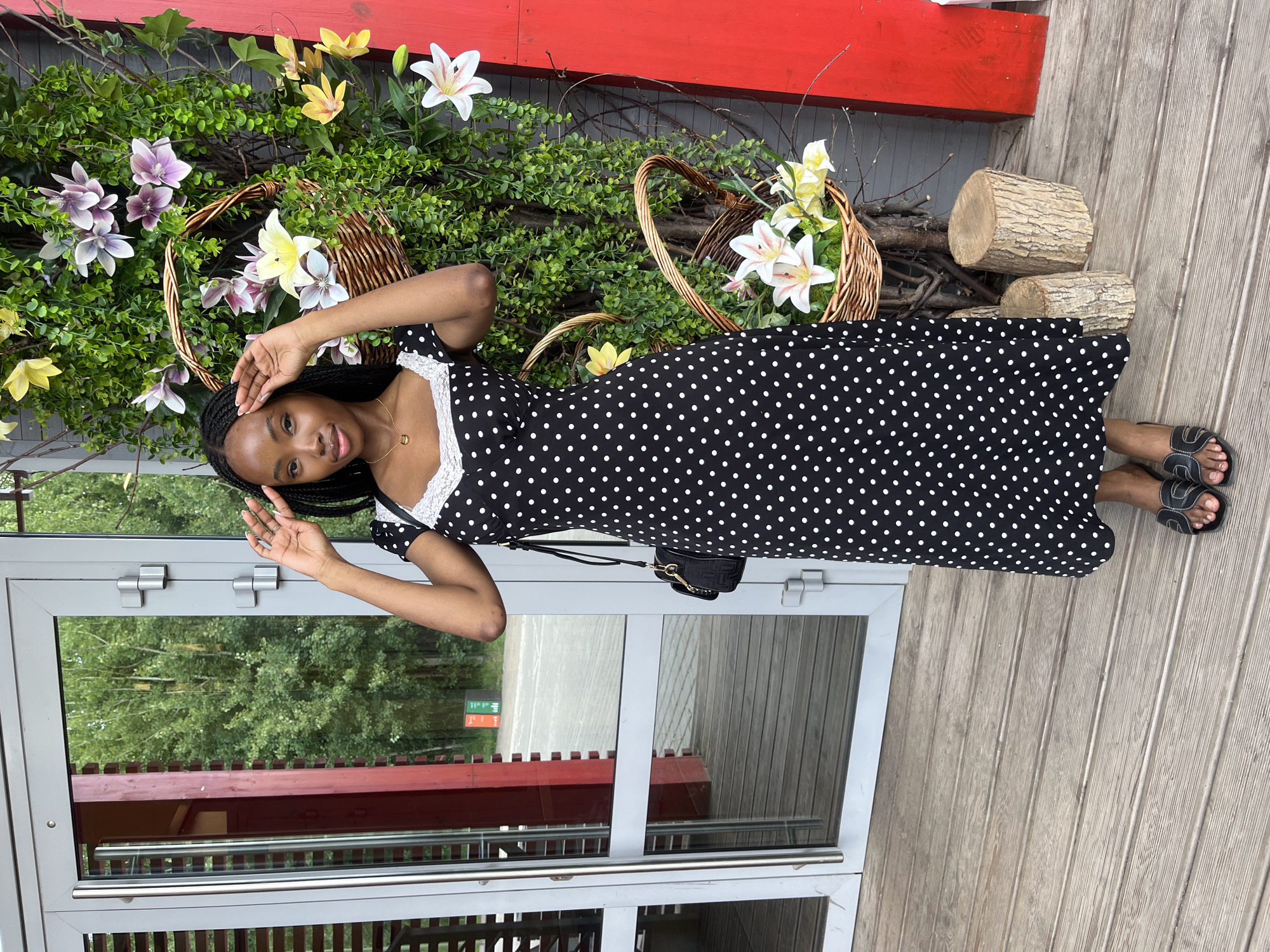 A young Black woman is standing on a wooden porch in front of a building with glass doors. She is wearing a black and white polka dot dress with white lace trim, and black sandals. She has her hands on her head, posing for the photo. Behind her is a decorative wall covered in greenery and artificial flowers, with two wicker baskets filled with more flowers. To her right are two tree stumps and a bright red vertical structure. The background visible through the glass doors shows trees and an outdoor area. This scene is located in the city of Kazan, Russia.