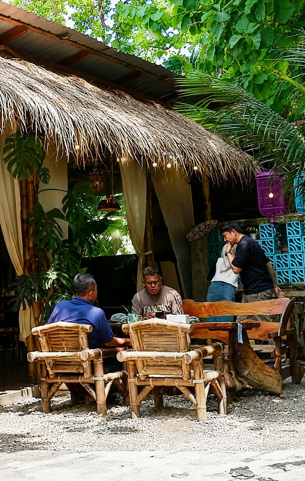 The image depicts an outdoor seating area of a restaurant or cafe, likely located in the City of Medan, Indonesia, given the tropical foliage and architectural style. The scene is characterized by a thatched roof structure with bamboo pillars and cream-colored drapes.
There are four individuals present. Two men are seated at a wooden table, facing away from the camera, engrossed in conversation. One man, wearing a blue shirt, is on the left, while the other, in a beige t-shirt with the word "FAMOUS" visible, is on the right. They are seated on woven bamboo chairs. In the background, to the right of the table, a man in a black t-shirt and a person in a white top and blue jeans appear to be interacting, with the man in black leaning towards the other person.
The seating area is paved with gravel. The environment is lush with greenery, including large-leafed plants and palm fronds visible above the thatched roof. Decorative elements such as string lights and a purple basket add to the ambiance. A wooden bench with carved detailing is also part of the seating arrangement. The overall impression is a casual, open-air dining space in a tropical setting.